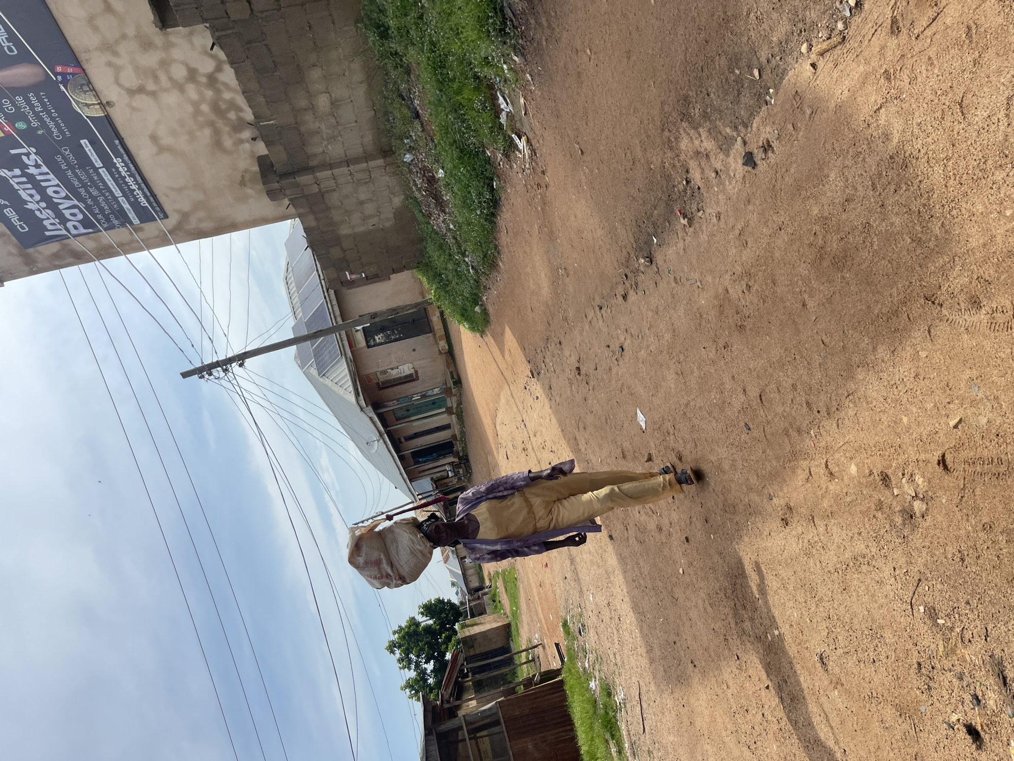 A woman is walking down a dirt road in Rafin Par, Nigeria, carrying a large sack of goods on her head. She is wearing a tan jumpsuit and a patterned jacket. The road is lined with buildings on both sides, some of which appear to be shops or homes. There are power lines overhead and solar panels on the roof of one of the buildings. The sky is cloudy. In the background, there is a large billboard advertising "Instant Payouts!" and "Crypto Trading". The billboard also lists phone numbers and a website address.