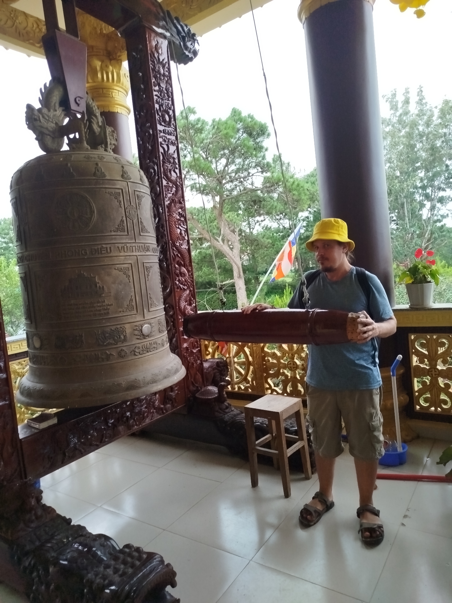 In Da Lat, Vietnam, a man wearing a yellow bucket hat, a blue t-shirt, and khaki shorts stands on a tiled floor. He is holding a large, wooden, cylindrical object, possibly a gong mallet, with both hands. To his left is a massive, ornate bronze bell, suspended by a dark wooden frame carved with dragon motifs. The bell is adorned with intricate patterns and script. In the background, behind the man, are trees, a building with ornate yellow and brown columns, and a flag with orange, white, and blue stripes. A small wooden stool and a potted plant with red flowers are also visible. The scene appears to be at a Buddhist temple or a cultural site.