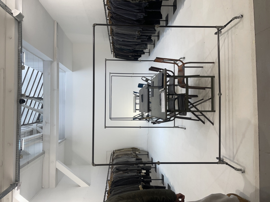The scene depicts a minimalist, brightly lit interior space in Paris, France, likely an exhibition hall, showroom, or studio. The environment features white walls, a white floor, and a white ceiling with exposed white-painted structural beams and visible skylights providing natural light. Overhead, artificial lighting fixtures and a security camera are mounted.
The room is organized with two long, black metal pipe garment racks lining the left and right sides, filled with numerous uniform-style jackets or coats in muted dark and earthy tones. In the central area, three progressively smaller rectangular structures, constructed from black metal pipes, are arranged in a receding line.
Within the foreground structure, a dark rectangular table is set with two black folding chairs to the left and one brown wooden chair to the right. A silver laptop is open on the right side of the table, beside what appears to be papers or a notepad. Additional similar tables and chairs are visible within the subsequent structures further back.
In the lower-left corner, a partial view of an individual is present, showing the left arm in a green jacket sleeve, and the lower body wearing light-colored trousers and brown footwear. The person is standing, facing towards the central arrangement. No specific actions or interactions beyond their presence are discernible.