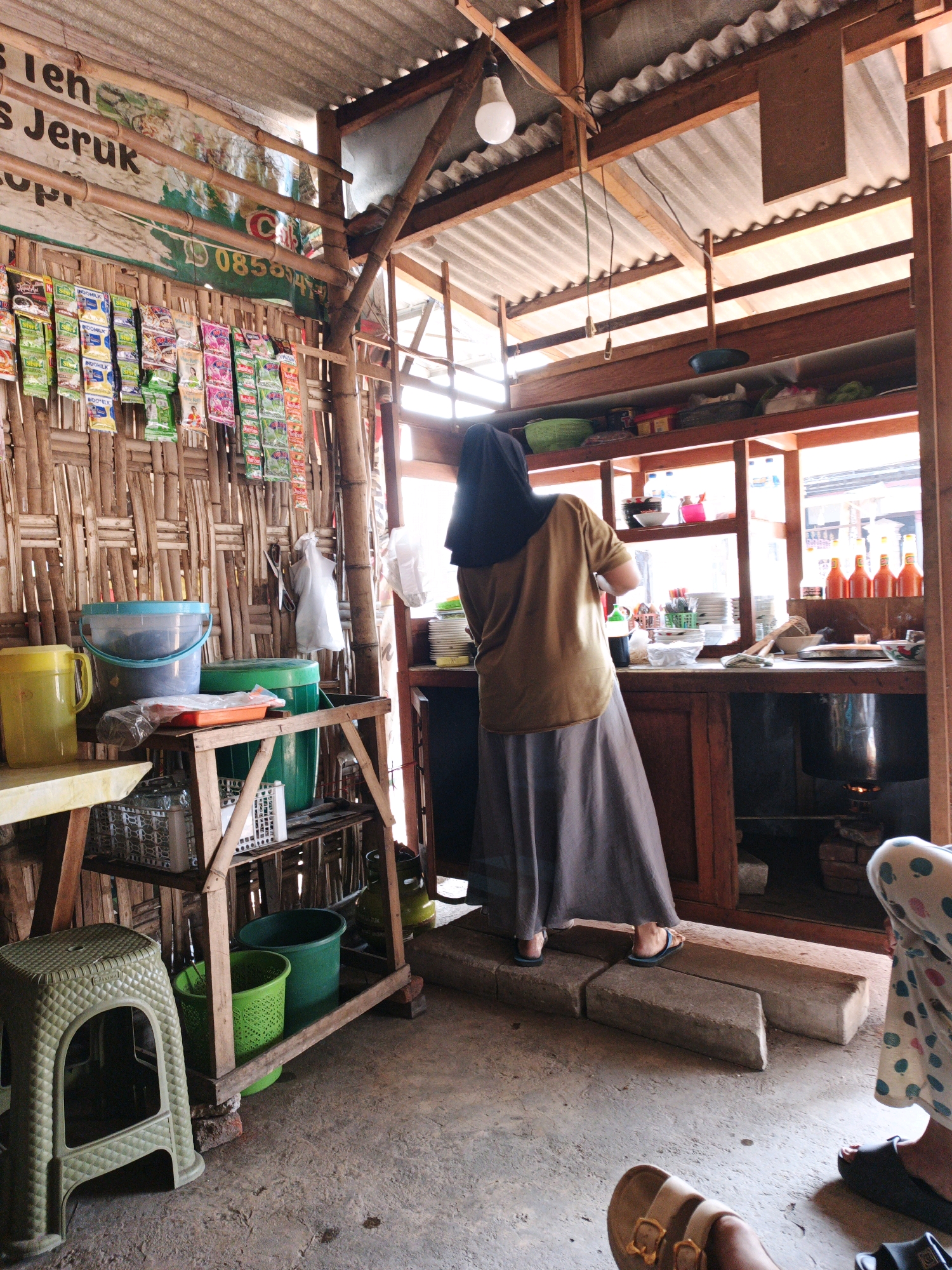 The image shows the interior of a small food stall in Wonorejo, Indonesia. The stall has a rustic appearance with bamboo walls and a corrugated metal roof. In the foreground, a green plastic stool is placed on the concrete floor. To the right of the stool, a wooden shelf unit holds various items, including buckets, plastic containers, and what appear to be groceries.
In the middle ground, a woman wearing a brown long-sleeved shirt and a long gray skirt is standing and appears to be working at a counter. She is also wearing a black headscarf. The counter is made of wood and has shelves above it, displaying plates, bottles, and other kitchen items. Various bottles of beverages and packets of food are hanging on the bamboo wall behind her.
In the background, through an open doorway, parts of the exterior of the stall are visible, suggesting it's a street-side establishment. The overall impression is of a simple, functional food preparation and service area.