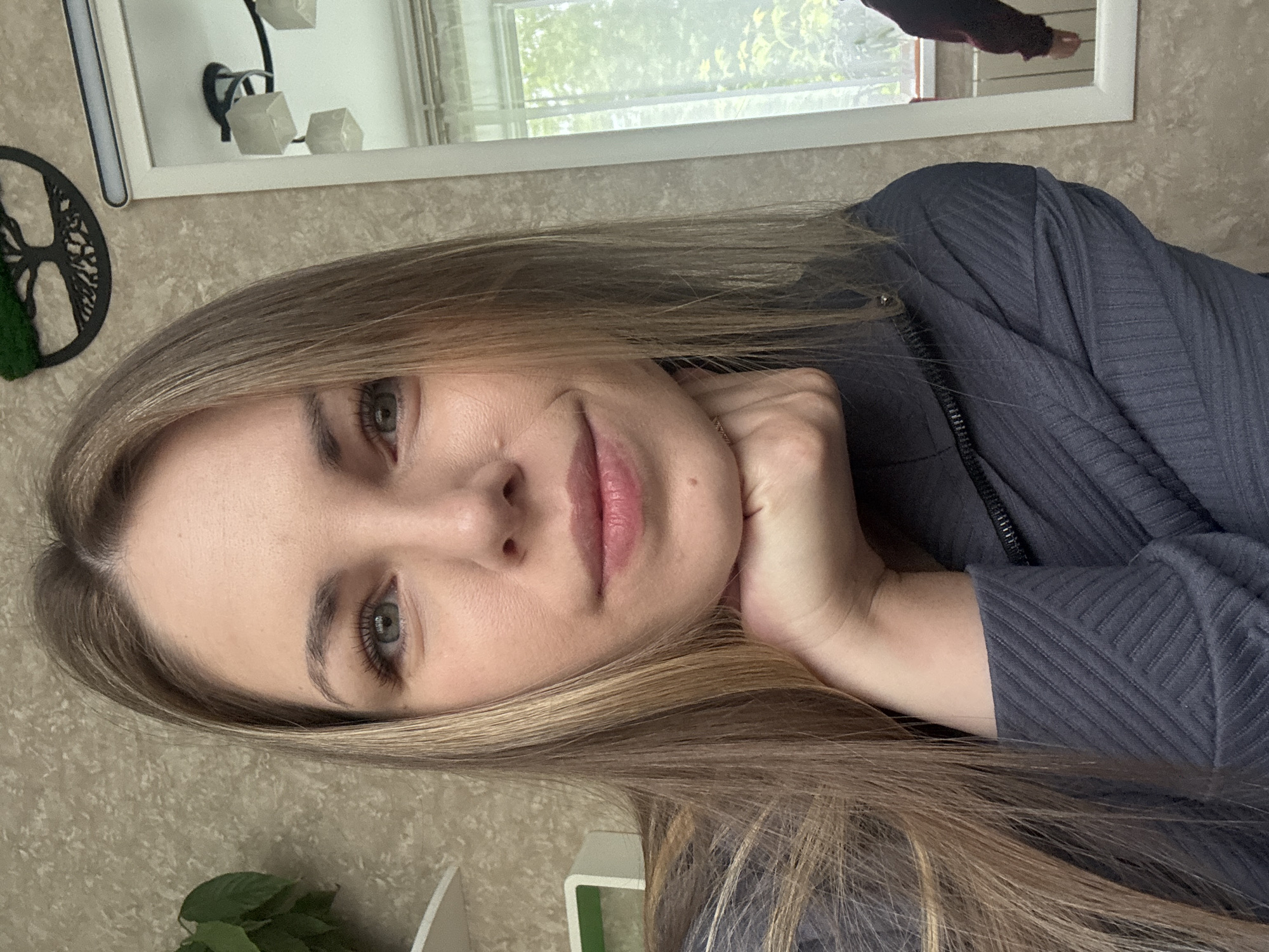 A woman with fair skin and light brown hair is resting her chin on her hand. She has grey eyes and pink lips. She is wearing a dark grey top with a ribbed texture. The background includes a beige wall, a white-framed mirror, and a window with a view of trees. There is also a decorative tree and a plant in the foreground. The reflection of a person's arm can be seen in the mirror. There is no evidence to connect the location to Раздолье, Russia.