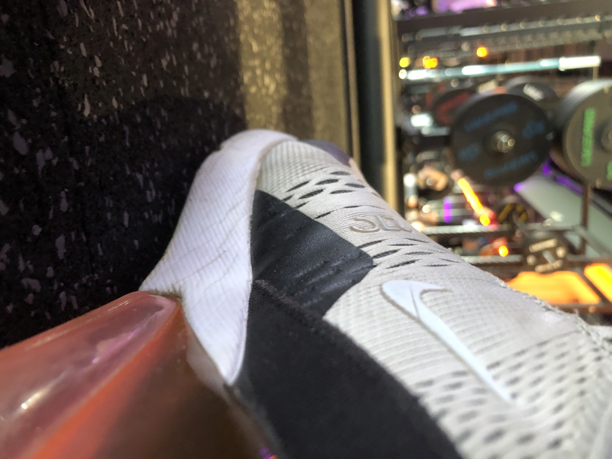 A close-up, angled shot focuses on a person's foot wearing a white, black, and light gray Nike Air Max 270 sneaker. The shoe's upper features a textured mesh material and a prominent white Nike swoosh logo on its side. The numerical "270" text is visible on the tongue or upper part of the shoe. The heel of the sneaker showcases a distinctive translucent orange-pink Air sole unit. The shoe rests on a dark, textured surface, likely a rubber gym floor or similar material.
In the blurred background, an indoor setting is suggested by an array of vibrant artificial lights in yellow, orange, blue, and purple hues, along with dark, circular objects that could be weights or components of a machine, possibly in a gym, arcade, or entertainment venue. The lighting conditions imply an evening or nighttime scene, or a dimly lit interior. No specific activity is depicted other than the shoe being stationary. There are no people visible other than the foot in the sneaker. The location is Krasnoyarsk, Russia.