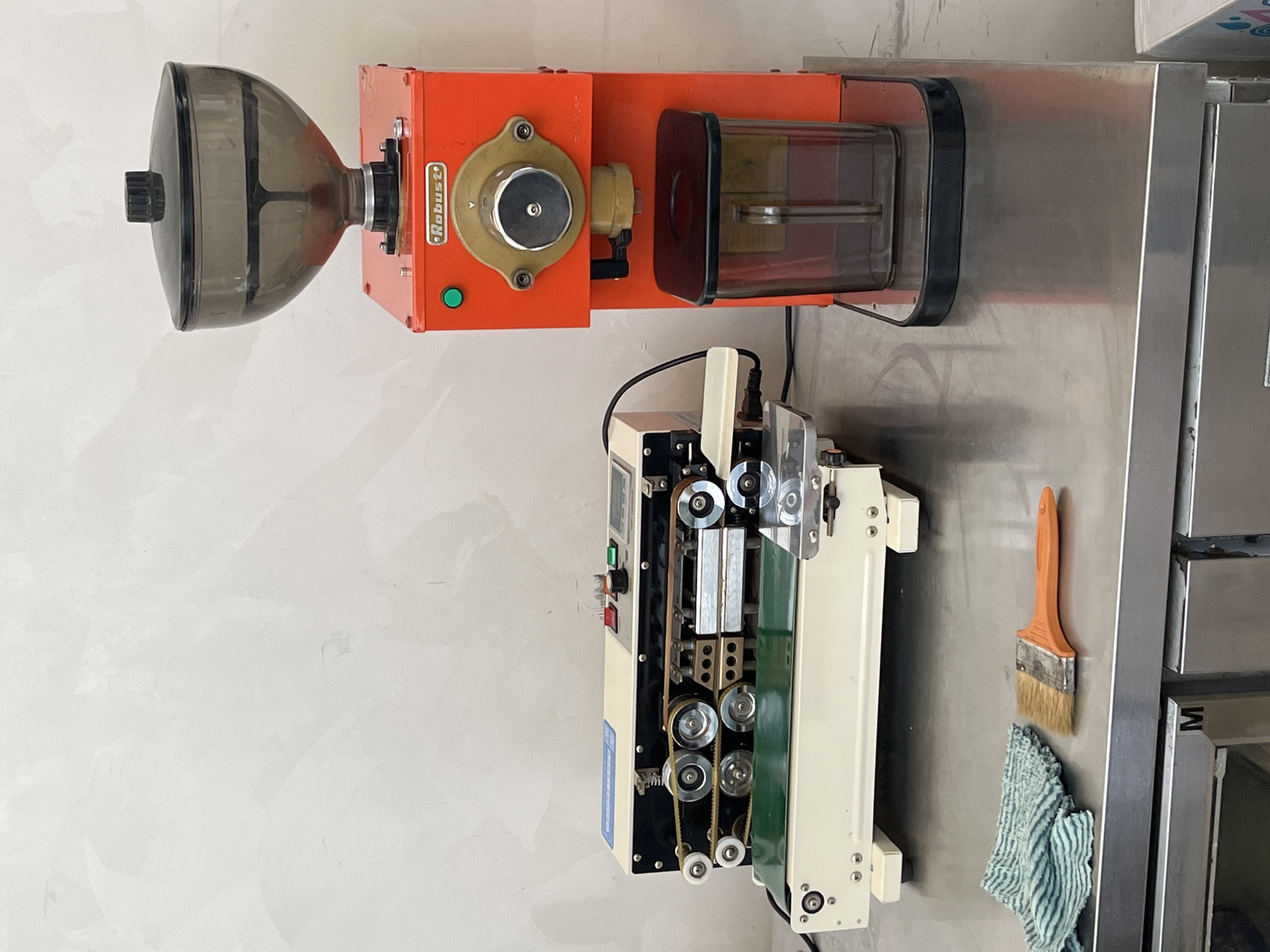 The image shows a commercial kitchen setup. At the top is an orange "Robust" branded coffee grinder with a clear plastic bean hopper. Below is a bag sealing machine with rollers, belts, and a control panel. Adjacent to the bag sealer is a stainless steel countertop. A brush and a striped cloth are placed against the steel. The location is Bac Nha Trang Ward, Vietnam, as indicated by the location context. No people are visible.