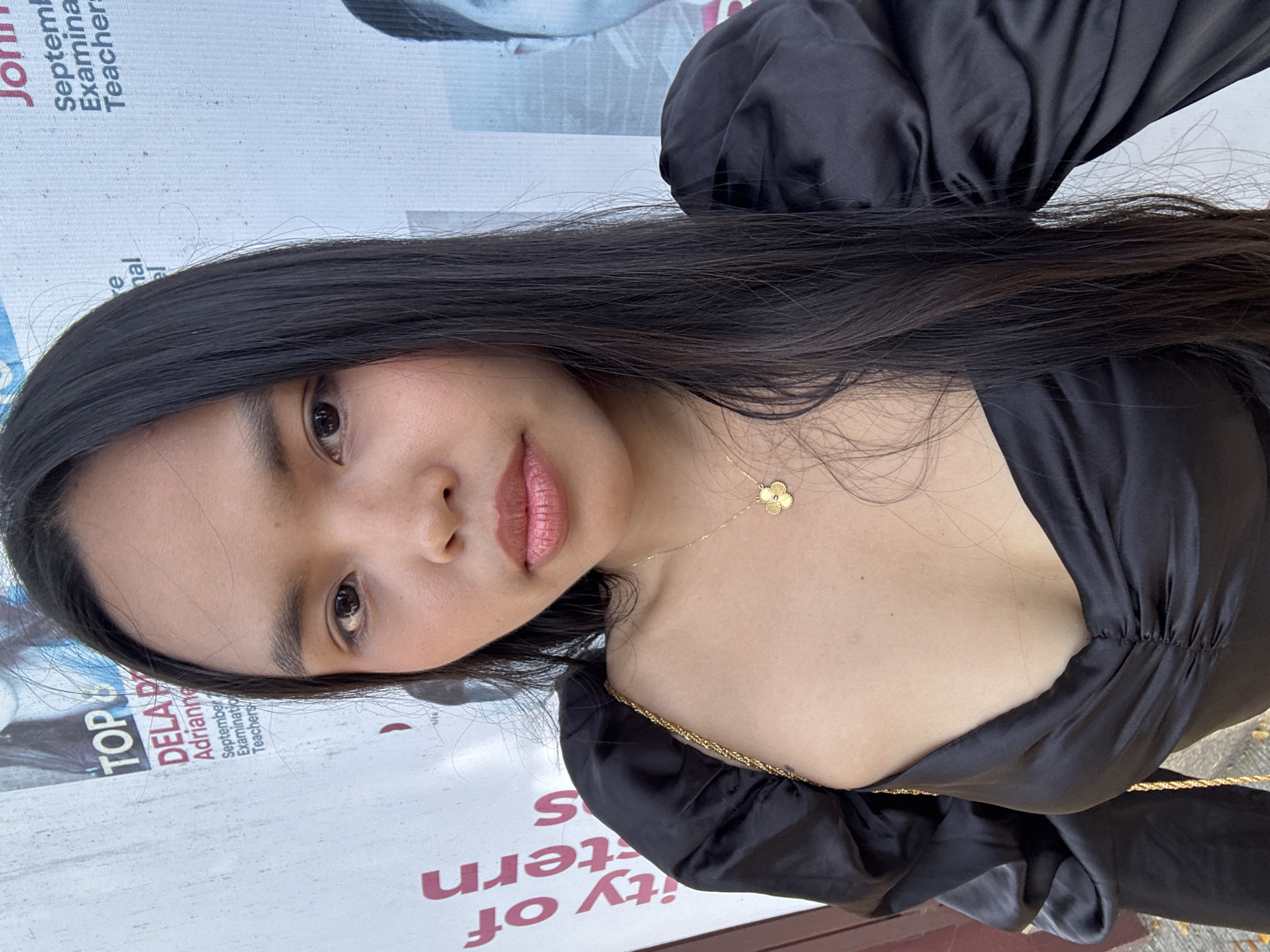 A young woman, appearing to be of Southeast Asian descent, is featured in a close-up, chest-up portrait, looking directly at the camera with a neutral, calm expression and slightly parted lips. Her head is tilted slightly to one side. She has long, straight, dark hair framing her face and falling over her shoulders. She is wearing a black top, likely made of satin or a similar silky material, featuring puffy sleeves and a V-neckline. A delicate gold chain necklace with a small, four-petal gold flower pendant adorns her chest, and a gold braided strap, possibly from a bag, is visible over her right shoulder. Her makeup includes a pinkish-brown lipstick and defined eyebrows.
The background is a light-colored surface, which appears to be a large poster or banner with text primarily in red and blue, oriented vertically. Visible text fragments include "TOP", "DELA P", "Adrianne", "September 2", "Examinatio", "Teachers", "Septemb", "nal", "Examina", and "Teacher:". There's also partially visible, rotated text that suggests "University of Western" or similar ("ity of ern S"). These texts indicate an educational context, likely an announcement for a teachers' examination or event scheduled for September. The lighting is bright, suggesting the photo was taken during the daytime in what appears to be a public or institutional setting in Davao City, Philippines.