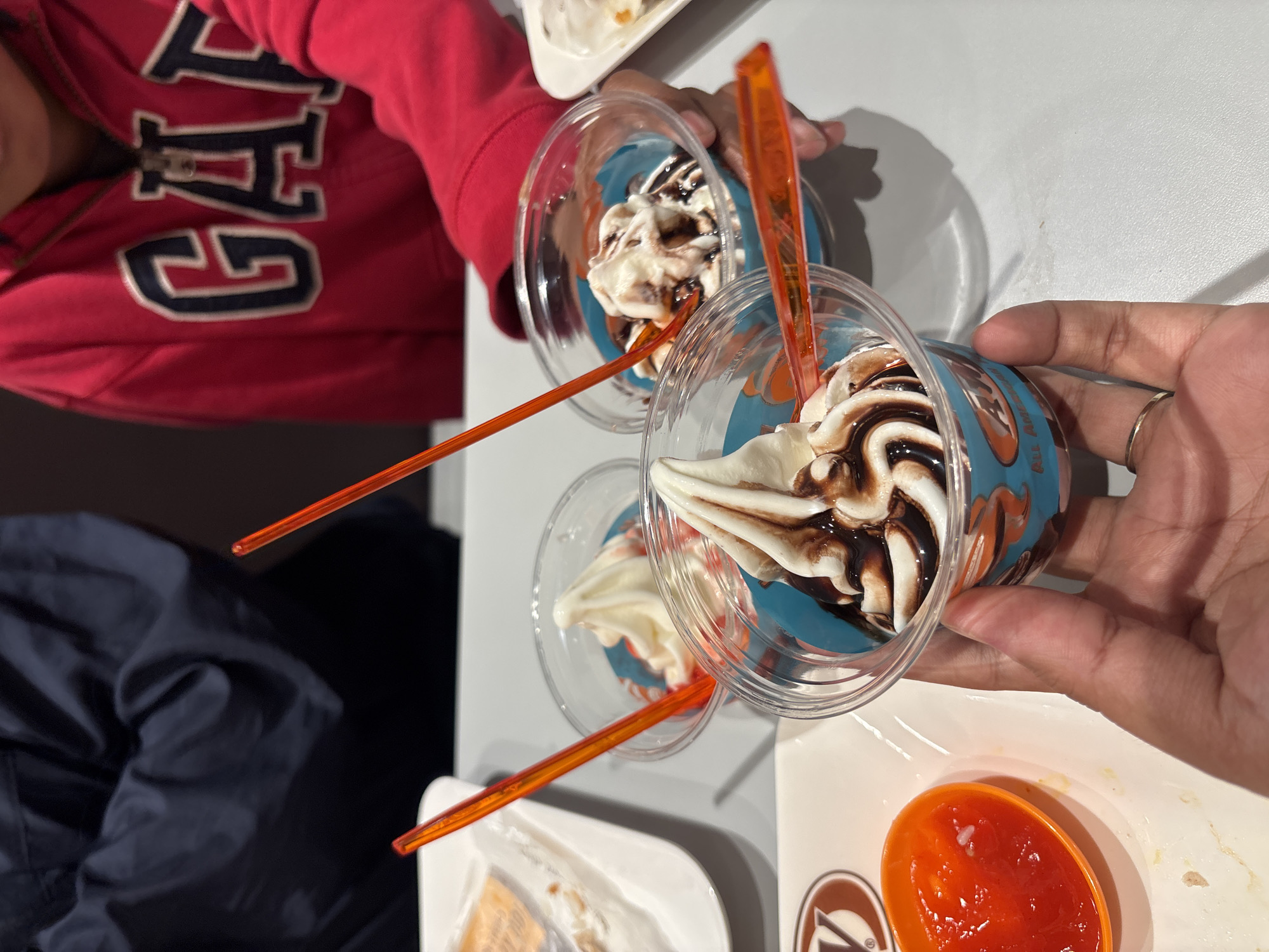 In Sukabakti, Indonesia, at a table, a person's hand, adorned with a silver ring, holds a clear plastic cup containing soft-serve ice cream drizzled with chocolate sauce. The ice cream is swirled and sits atop a blue-colored base within the cup, which has a printed design featuring orange and blue motifs. An orange plastic spoon is inserted into the ice cream. To the left, another cup of ice cream is visible, this one appearing to be plain vanilla. In the background, a person wearing a dark blue jacket and another person wearing a red hoodie with a "GAP" logo are partially visible, suggesting they are at the same table. A small orange bowl filled with a red sauce, possibly chili or fruit-based, sits to the lower left of the frame on a white tray. The setting appears to be a casual dining establishment.