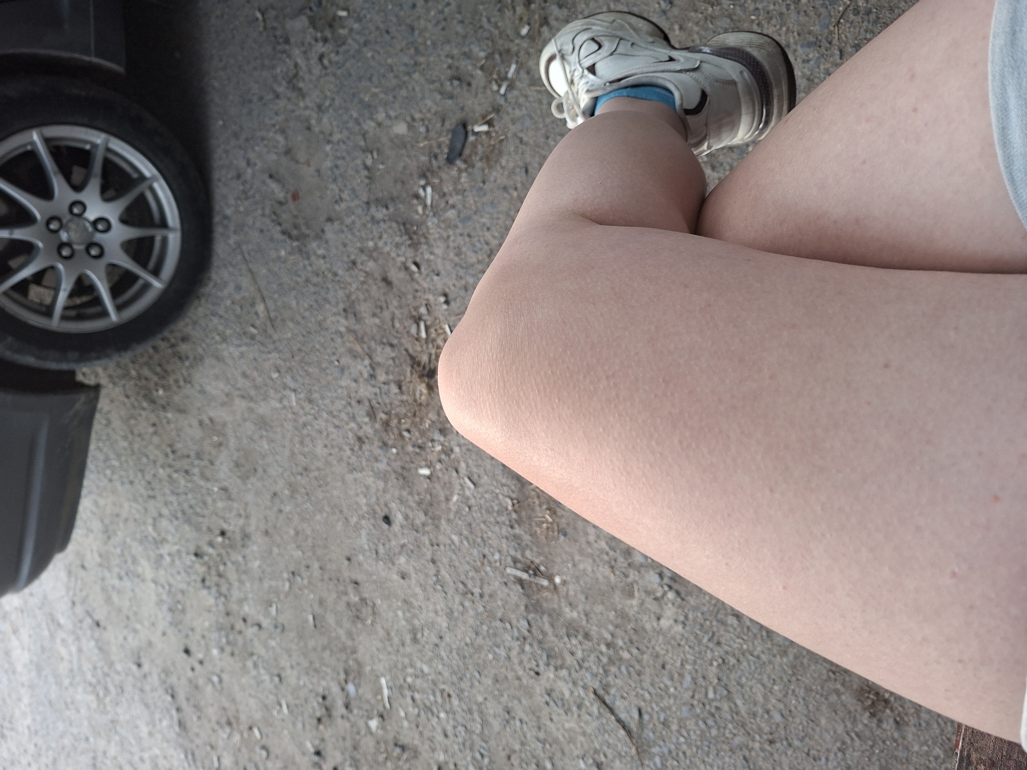 The image shows a partial view of a person sitting outdoors, with crossed legs, wearing a white sneaker and a blue sock. A car wheel is visible on the left side. The ground appears to be concrete or gravel. The location is Galabovo, Bulgaria.