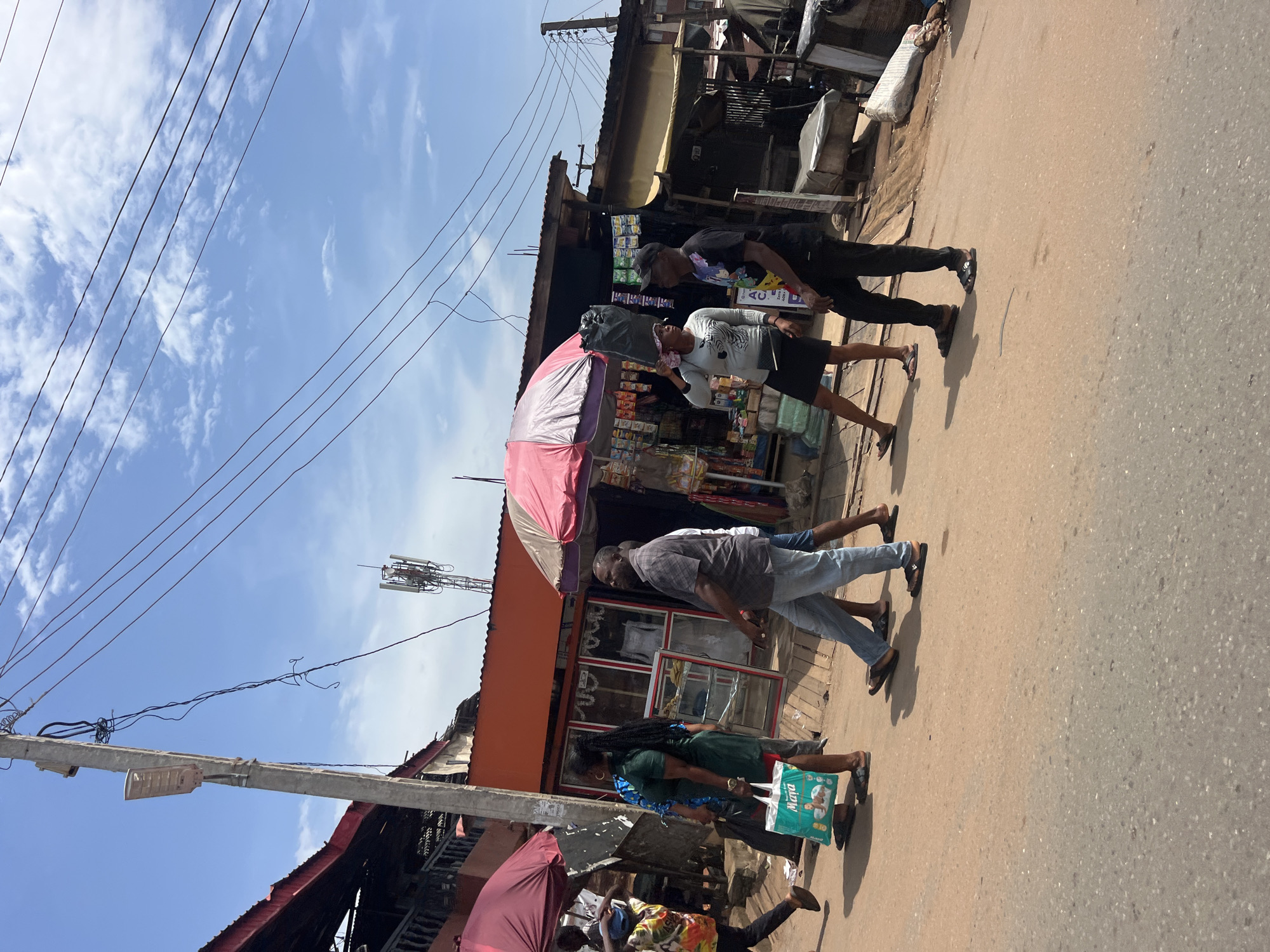 In Nkpor, Nigeria, a street scene unfolds with several individuals walking along a dusty, paved road. The environment features a row of market stalls or shops lining the right side of the road, some with open fronts displaying goods. Overhead, a network of power lines crisscrosses the sky, punctuated by a utility pole with a street lamp and a distant cell tower.
On the left side of the road, market umbrellas, one a deep red and another a pink and white striped one, offer shade. People are seen walking in both directions. A woman in a green dress carries a light blue shopping bag with the word "Maya" visible. Another woman in a black skirt and light-colored patterned top carries a large black plastic bag over her head. A man in a checkered shirt walks by, while another man in a black t-shirt and black cap is seen talking to the woman carrying the bag. Further down the road, a person in a colorful patterned shirt and black pants walks away from the camera. The ground is a mix of packed earth and asphalt. The sky is bright blue with scattered white clouds.