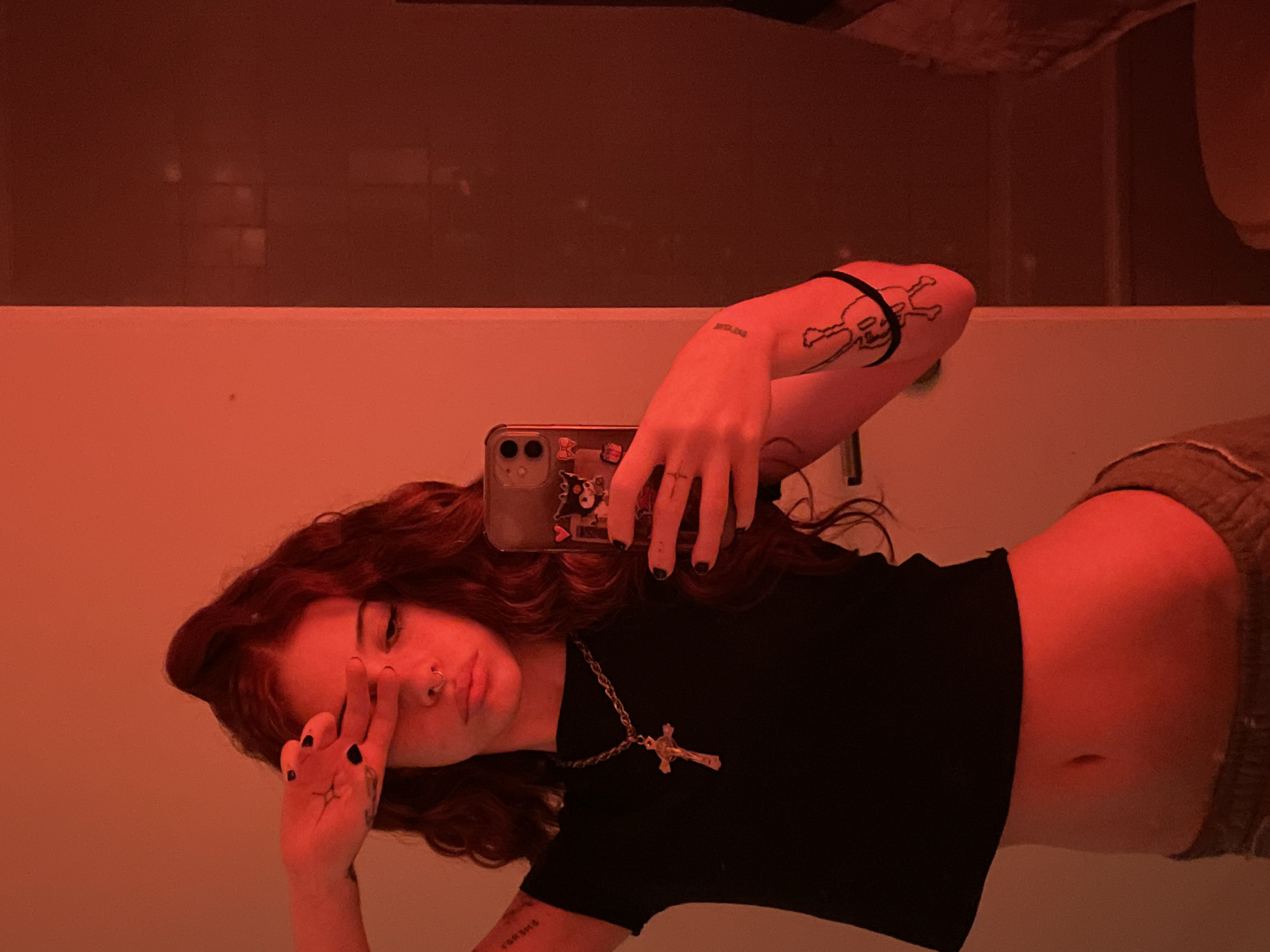 An indoor scene, illuminated by strong red-orange lighting, depicts a young female subject taking a mirror selfie. The subject, positioned centrally, has long, wavy, dark hair, styled partially swept up. She wears a black cropped t-shirt, exposing her midriff, and light-colored sweatpants. A silver-colored chain with a crucifix pendant is worn around her neck. A septum piercing is visible on her nose. Black nail polish is present on her fingernails. Her left hand is raised to her forehead, fingers spread, partially obscuring her left eye, while her right hand holds a smartphone, angled to capture the reflection. The smartphone, consistent with an iPhone model, features a decorated case with stickers, including a cartoon character. Tattoos are visible on her left arm: text on the inner forearm and an outline design, possibly a cartoon figure or instrument, on the outer upper arm. A black elastic band is on her left wrist. The environment consists of a light-colored wall on the left, rendered reddish by the lighting, and a darker, reflective surface (likely a mirror) on the right, which shows a darker, indistinct background. No identifiable external markers or specific architectural features within the image indicate a precise location within Buenos Aires, Argentina; the setting is an interior room.