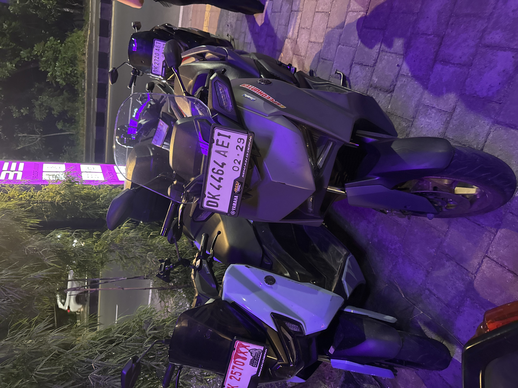The image depicts a row of parked motorcycles in Kuta Selatan, Indonesia. The foreground features a grey Yamaha motorcycle with the license plate "DK 4464 AEI," expiring on February 2029. Adjacent to it, partially obscured, is another motorcycle with a blue and black color scheme, its license plate reading "DK 2570XXX." Behind these, another motorcycle has the license plate "DK 2720 AET." The bikes are parked on a paved walkway illuminated by purple lighting. The lighting casts shadows across the walkway and the motorcycles. In the background, there is a blurry view of a street with a vehicle, and some greenery.