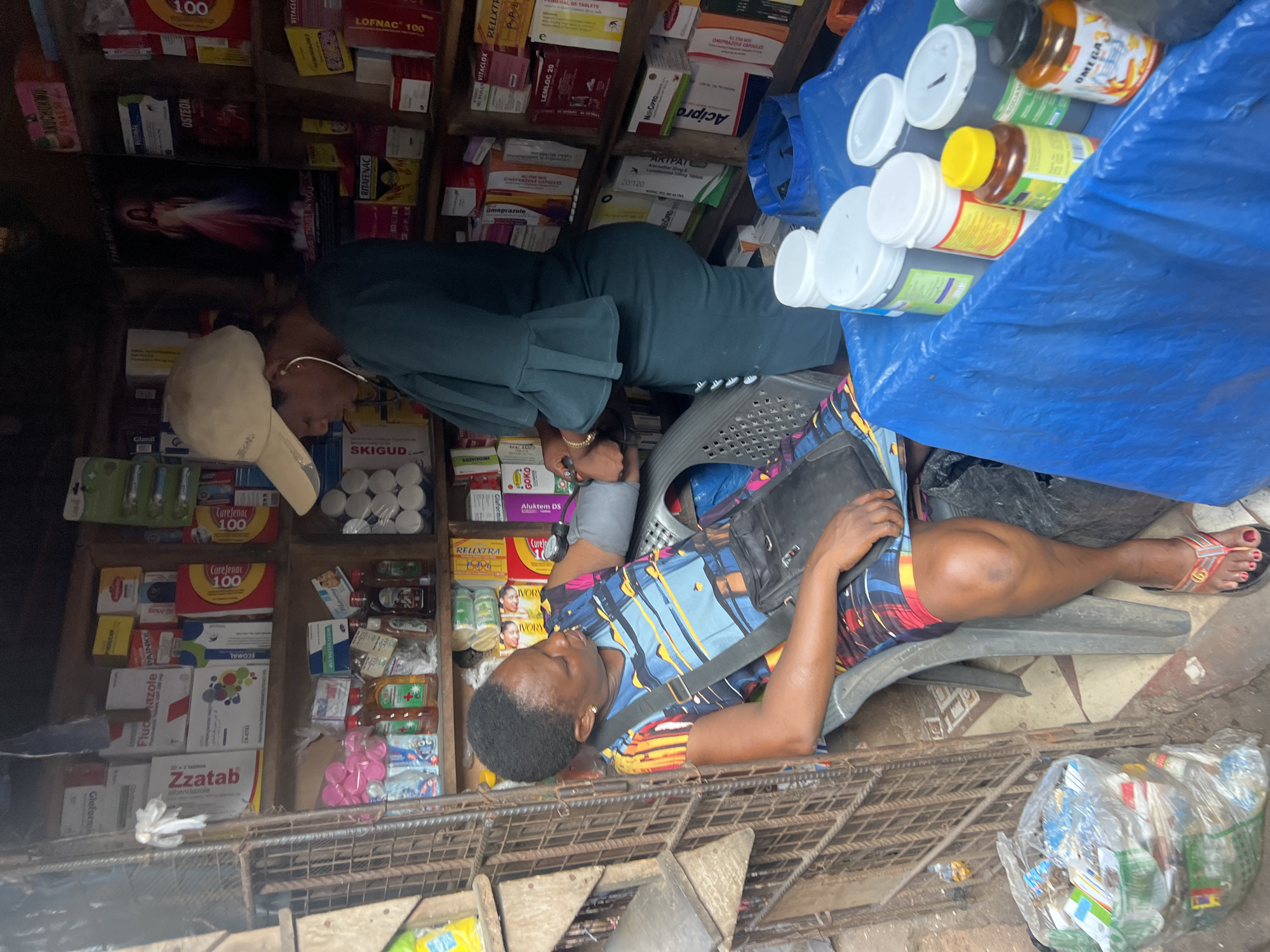 A street-level view depicts an open-front shop or kiosk, heavily stocked with pharmaceutical products. The shelves are laden with boxes and bottles of medications, various brands and types being prominently displayed. In the foreground, a woman in a colorful, patterned dress reclines on a chair, a black bag slung across her body. Beside her, there is a bag of assorted trash. Another woman, seen from the back, is bent over, seemingly examining items on the shelves. The shop appears to be in the city of Unknown, Unknown, based on the OCR text and the general setting. In the upper right corner, there is a blue tarp covering a stack of containers and jars.