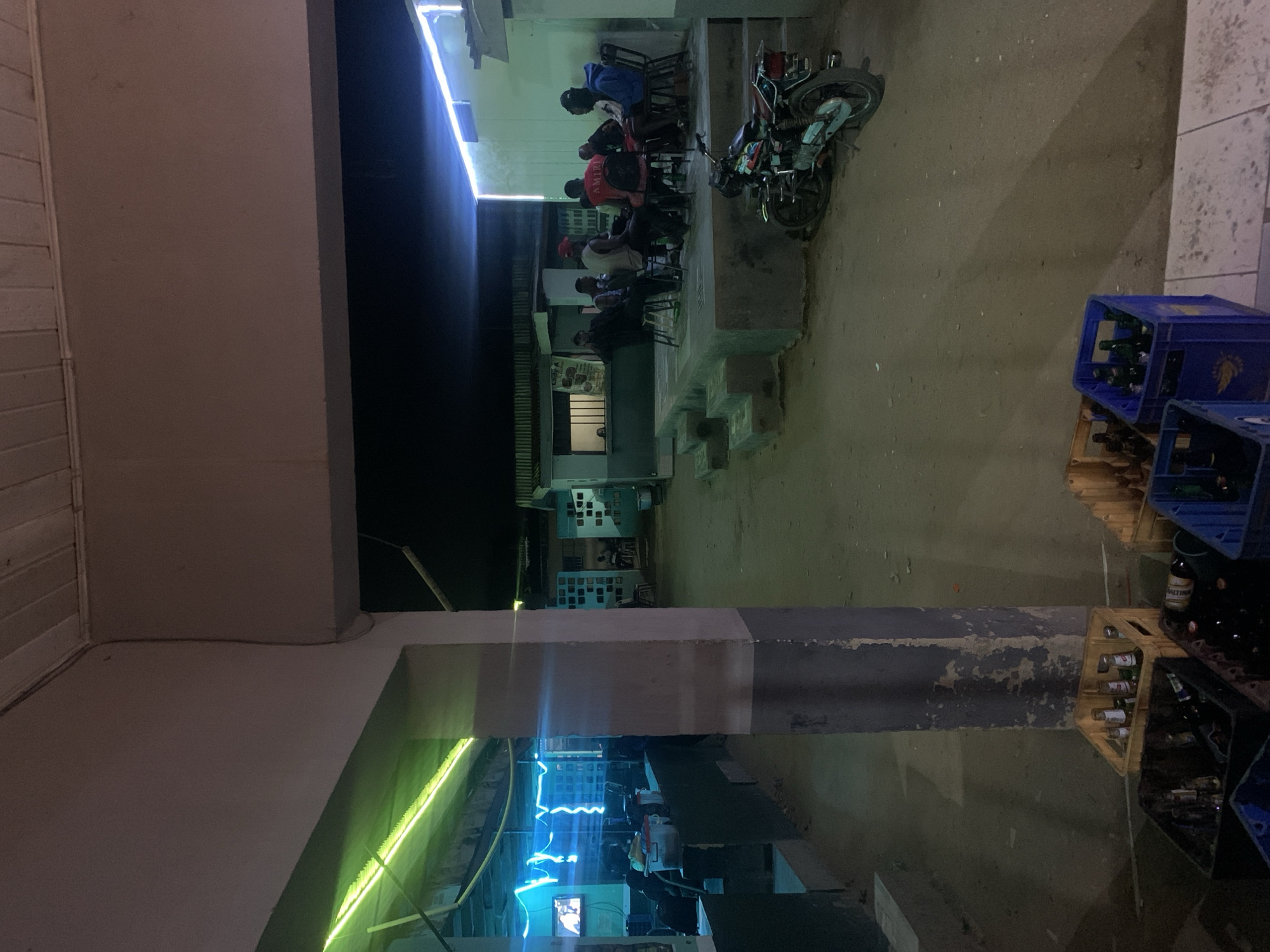 A nighttime scene in the city of New Stadium, Nigeria, shows a group of people seated at tables under a building overhang illuminated by white light. To the right, a motorcycle is parked. Blue crates containing bottles are stacked on a pallet. Below, more crates and a green light are visible.