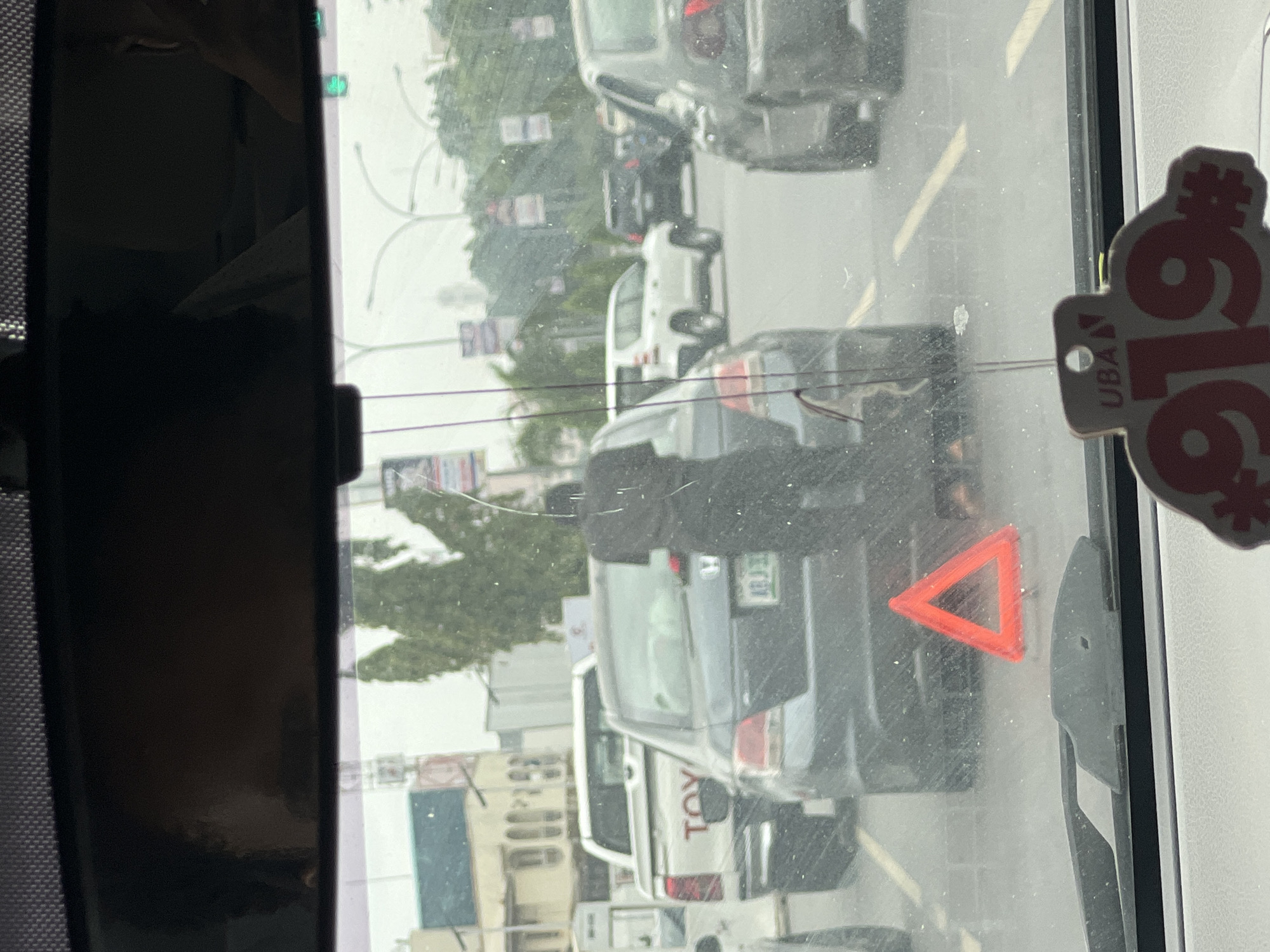 The image captures a daytime traffic scene in Abuja, Nigeria, viewed from inside a vehicle through a slightly dirty windshield. The perspective is from a stationary or slow-moving car, looking forward into a line of traffic.
On the left side of the frame, a black rearview mirror reflects a faint outline of a person, likely the driver or a passenger. On the lower right of the windshield, a bright red triangular hazard symbol is prominently displayed, suggesting the vehicle might be stopped or in distress. Hanging from the upper right corner of the windshield is a decorative or promotional tag.
Beyond the windshield, the road is filled with vehicles. Directly in front is a dark grey SUV or sedan, its rear lights and a partially visible license plate in view. Behind it, a white pickup truck, identifiable by the visible "TOY" text on its tailgate (indicating Toyota), follows. Further down the road, more cars are visible in the distance, suggesting a traffic jam or slow-moving queue. A green traffic light can be faintly seen in the distance, implying active traffic control. Buildings and scattered trees line the urban background under what appears to be an overcast or hazy sky.
The hanging tag on the windshield clearly shows the logo "UBA" (United Bank for Africa) and the prominent numbers "#616", likely a USSD code for mobile banking services. The overall scene depicts a typical busy road environment in a Nigerian city.
