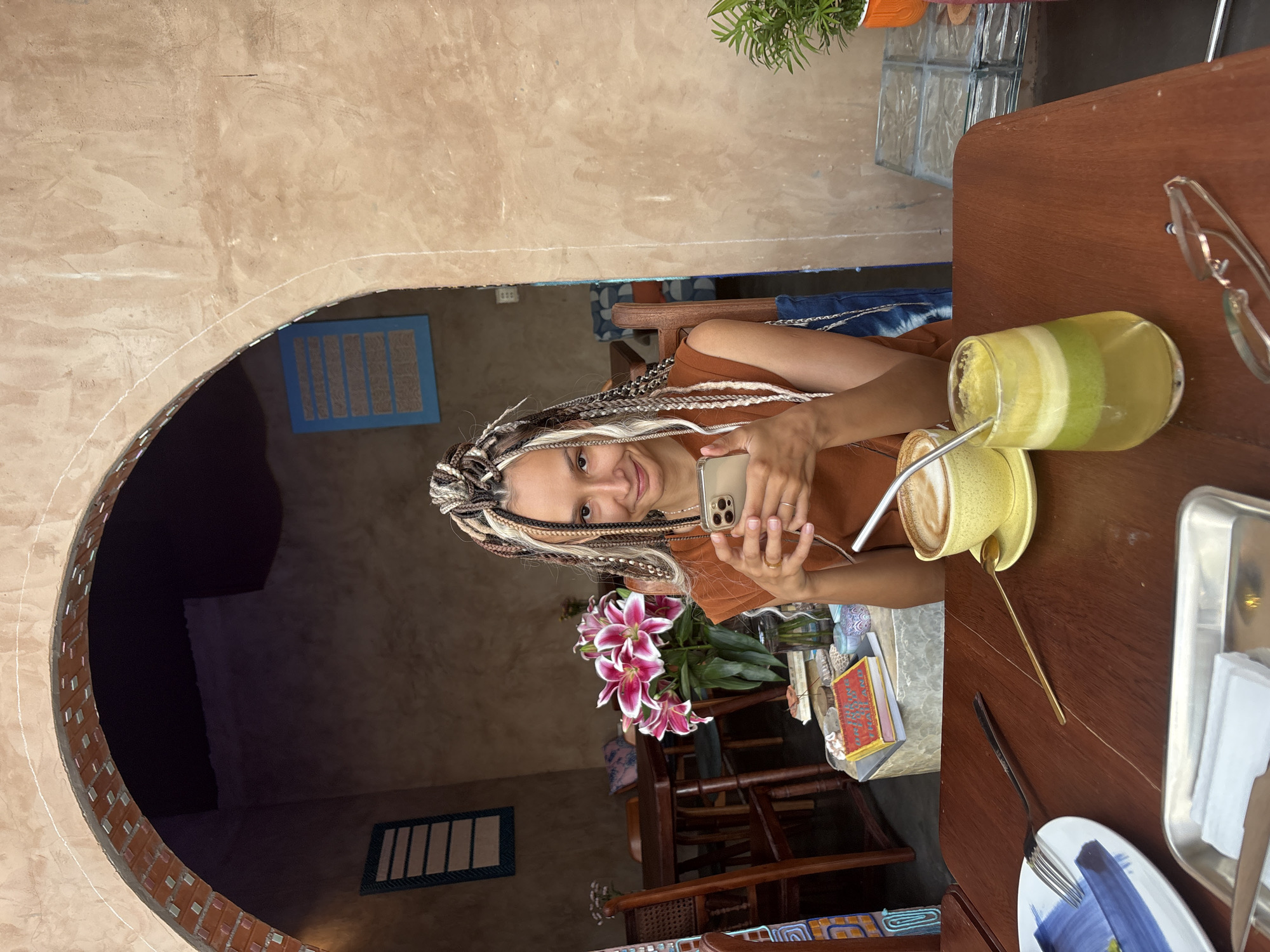 A woman with braided hair sits at a wooden table in a cafe in Mũi Né, Vietnam. She is holding a smartphone and looking at it. In front of her are a cup of coffee, a glass of juice, and a plate with a fork and knife. The cafe has a rustic, earthy interior with exposed brick and concrete walls. There are tables and chairs visible in the background, as well as some plants and decorations.