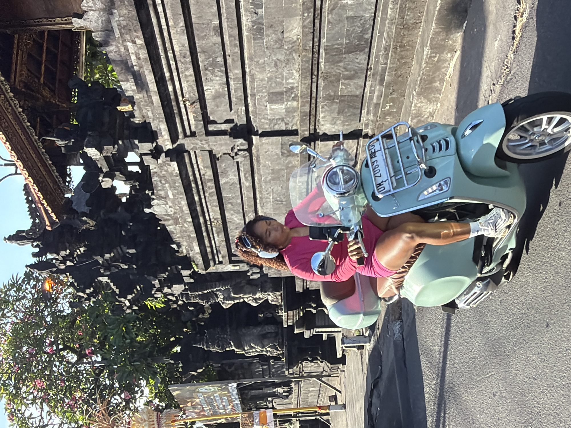 A woman with curly brown hair is riding a light blue Vespa scooter down a paved street in North Kuta, Indonesia. She is wearing a bright pink long-sleeved top and matching shorts, white headphones, and white athletic shoes. The scooter has a clear windshield, a chrome front rack, and a license plate that reads "DK 2069 AEQ". The background features a traditional Balinese stone wall with intricate carvings and sculptures, partially obscured by lush green trees with pink flowers. The sun is casting shadows on the ground, indicating it is daytime.