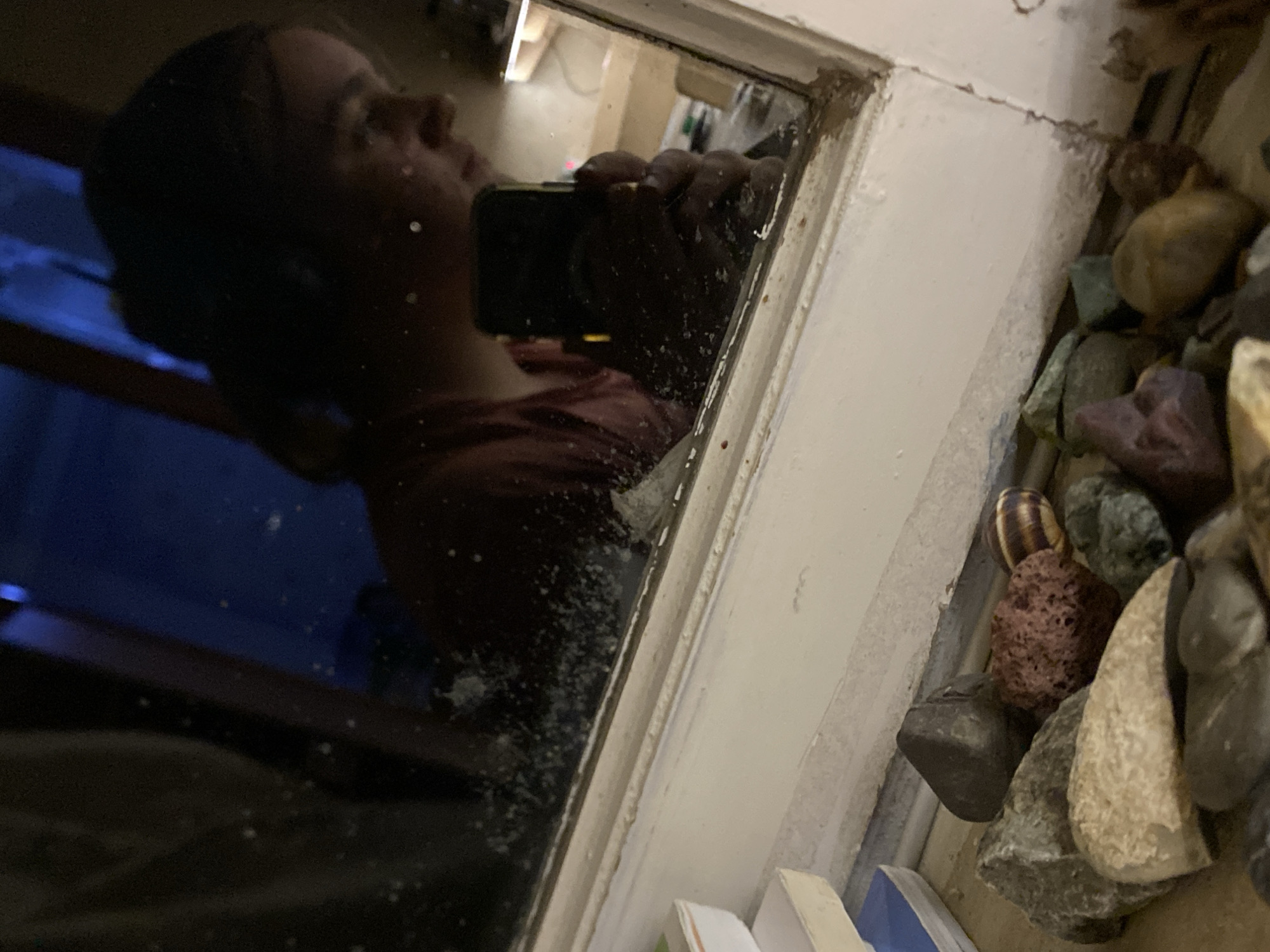 The image shows a reflection in a dirty window. The reflection includes a person holding a phone taking the picture. Outside the window, there is a collection of rocks and pebbles of varying colors and textures. The window frame is painted white and appears old and worn. The location, Tbilisi, Georgia, is not discernible from the image.