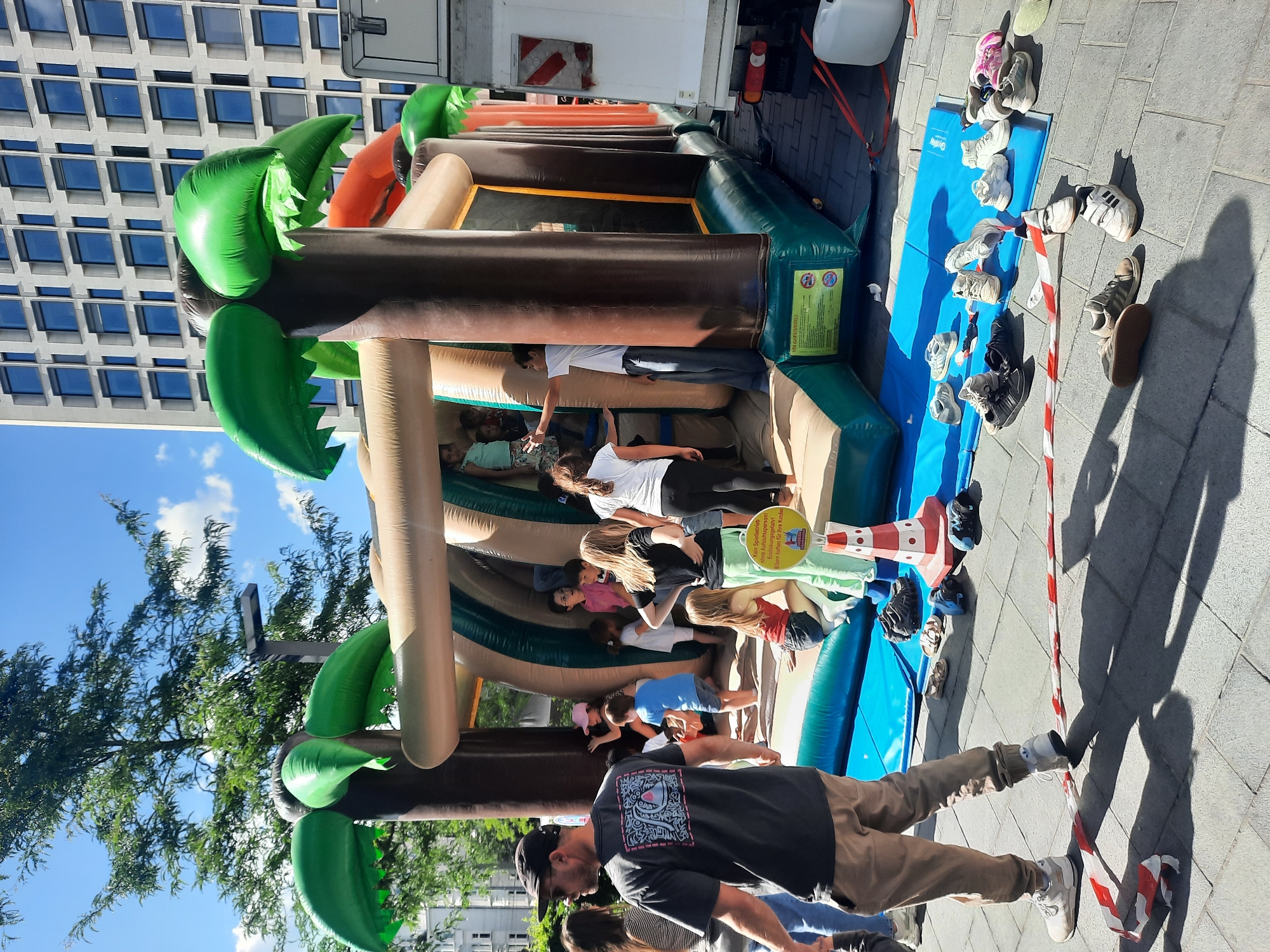 In an urban outdoor setting, an inflatable jungle-themed bounce house is set up on a blue mat on a tiled ground. The bounce house features brown palm tree trunks with green palm fronds and tan-colored tunnels. Several children are visible inside and around the bounce house. A man wearing a black cap and sunglasses stands to the left, facing away from the camera, with his back to the bounce house. To the right, a white truck is parked adjacent to the bounce house. Scattered on the ground are numerous pairs of children's shoes. In the background, a modern building with a grid of windows and a tree with green foliage are present. The sky is blue with scattered white clouds. A red and white striped caution tape is stretched across the ground in the foreground.