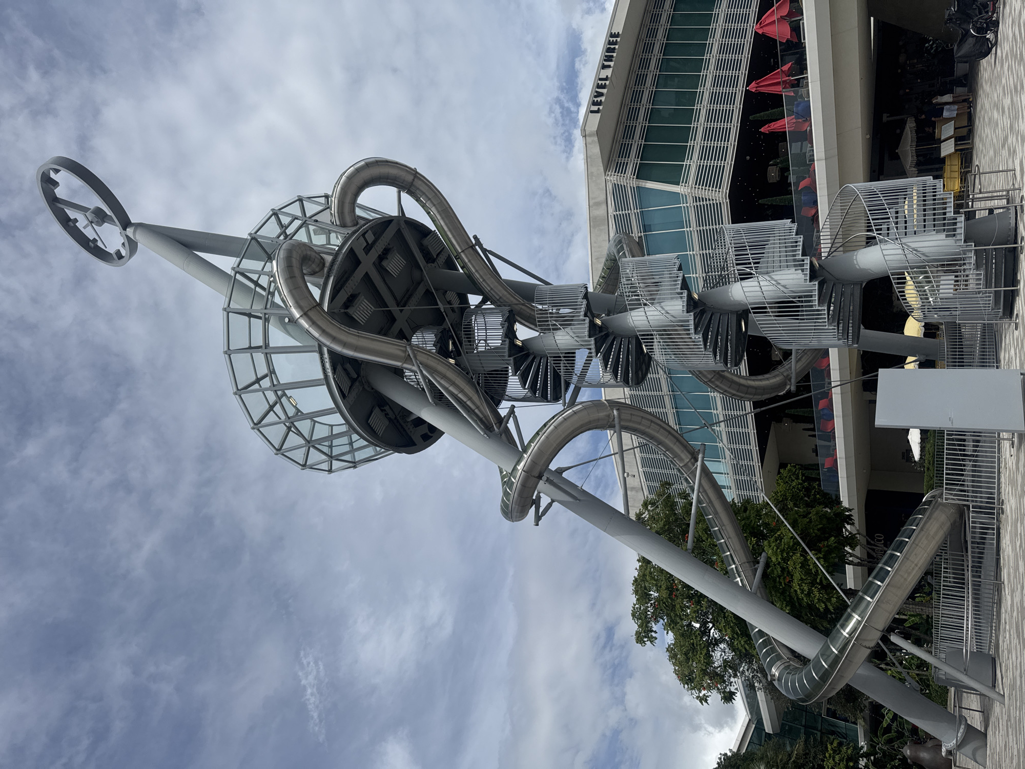 An outdoor shot features a large, elaborate metal slide structure with multiple winding tubes and spiral staircases. The structure is gray and made of metal, with some sections appearing to be made of glass or transparent material. It is set against a backdrop of a building with a sign that reads "LEVEL THREE." The building has large windows and is modern in design. Trees with green and orange foliage are visible to the left of the slide structure, and a cloudy sky fills the upper portion of the image. In the foreground, there are metal railings and a paved area. Red umbrellas are visible on a balcony of the building. The scene appears to be in a public outdoor space, possibly a park or a commercial area.