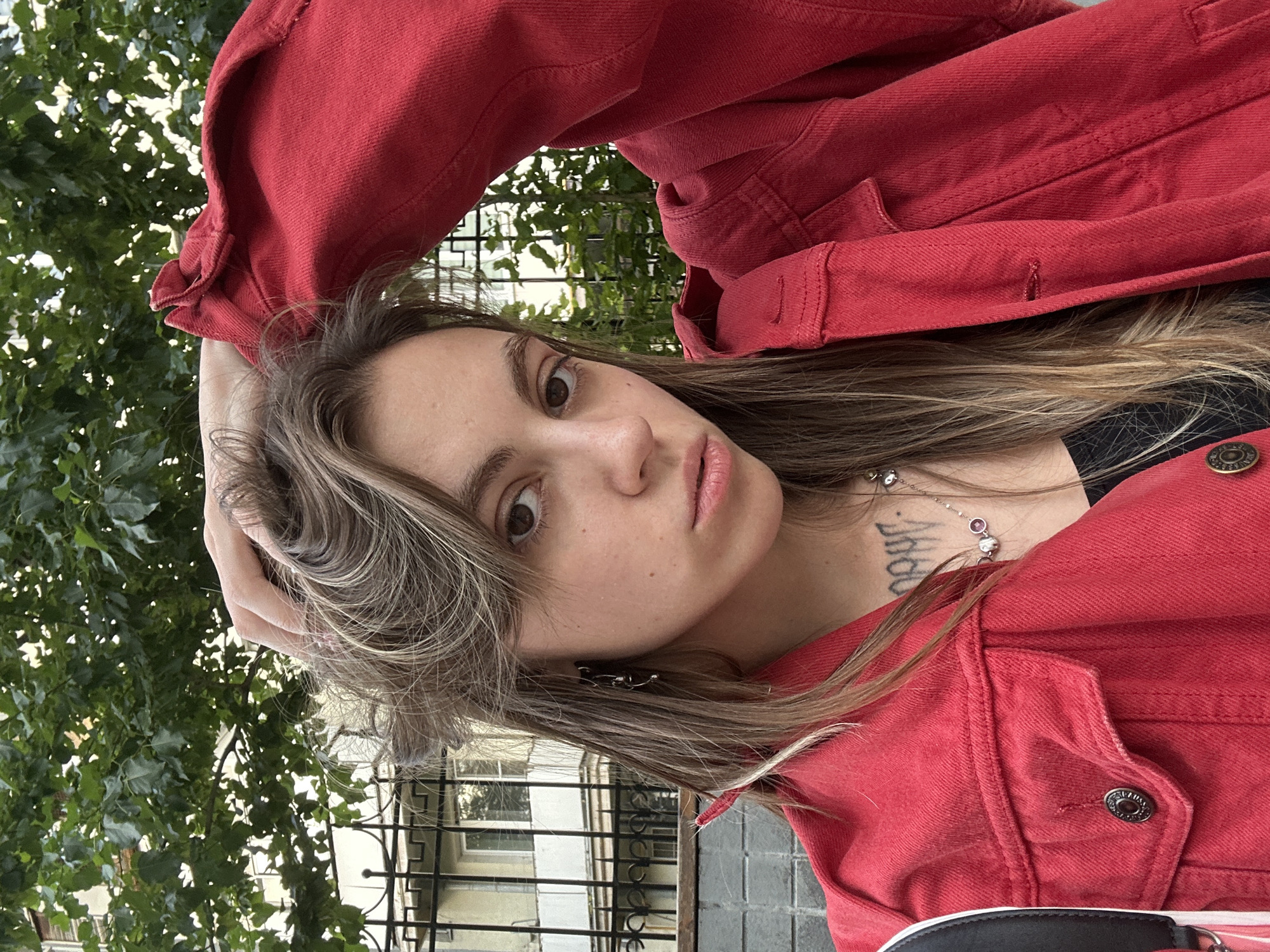 A young woman with long, blonde-brown hair, partially styled in bangs, is captured in a close-up portrait. She is wearing a vibrant red denim jacket over a black garment, and a delicate silver necklace with colorful beads adorns her neck. A visible tattoo with Gothic-style script is present on her upper chest. Her right hand is raised, touching her hair. The background suggests an urban environment, with lush green foliage from a tree in the upper portion of the frame, and a building with windows and a black metal fence visible behind her. The lighting indicates it is daytime.