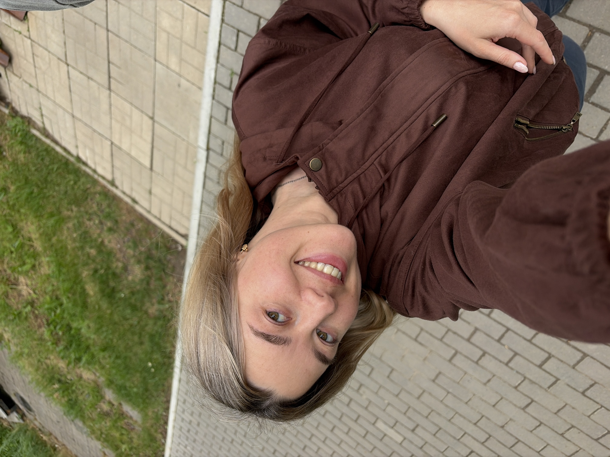 A woman with blond hair and a brown jacket is taking a selfie outdoors in Chaikovsky, Russia. The background includes a brick walkway, a patch of green grass, and a building. The woman is smiling and looking directly at the camera.