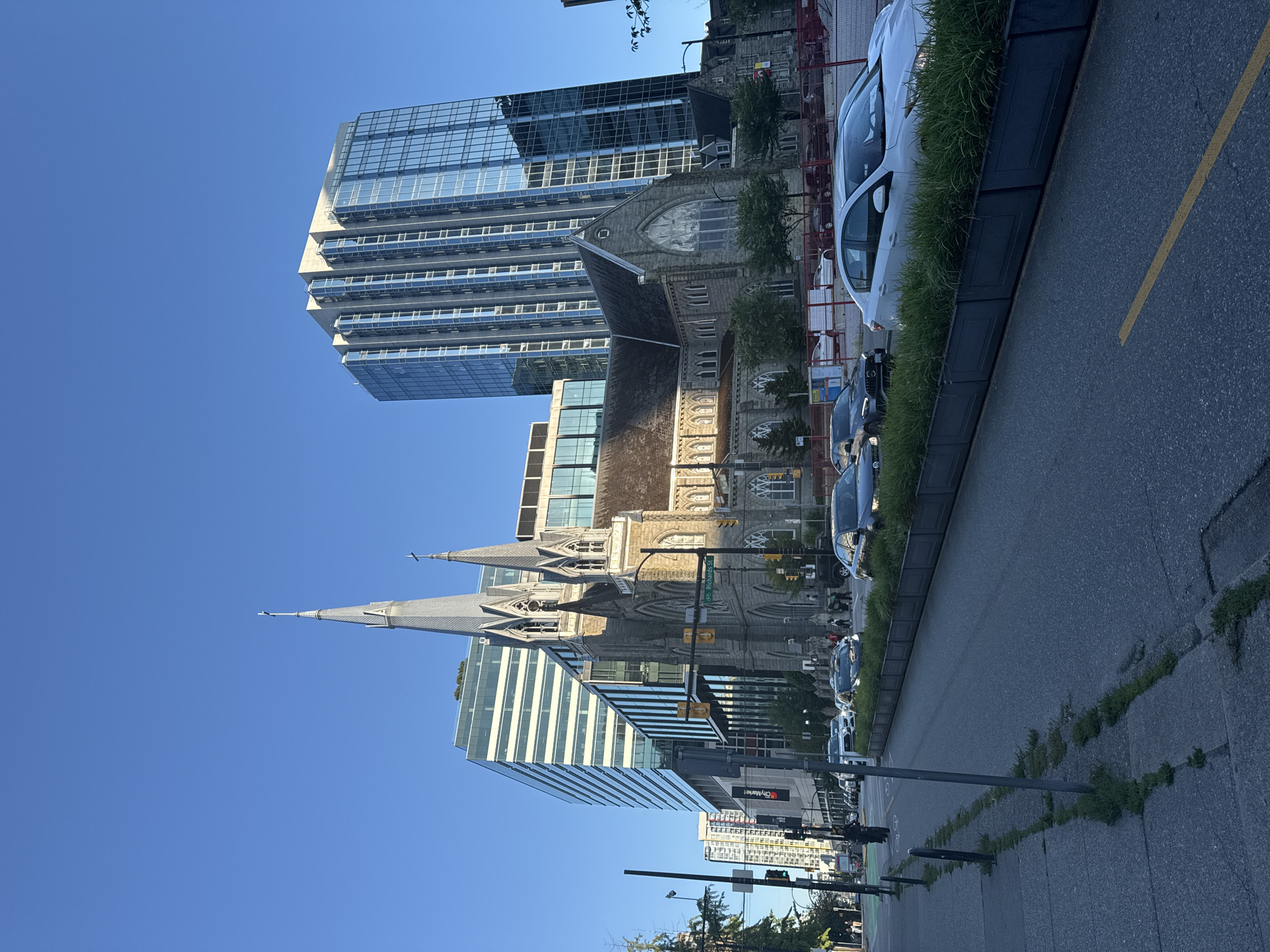 This image depicts an urban street scene in Vancouver, Canada, at what appears to be late afternoon or early evening, given the long shadows and clear blue sky. The focal point is a Gothic Revival church with two prominent spires, made of stone. Immediately behind and to the left of the church are modern, multi-story glass and steel buildings, one of which is particularly tall and sleek. To the right of the church, a smaller stone building is visible.
The street in the foreground is paved and shows lane markings, including a yellow line. A sidewalk runs along the left side of the street, with a metal pole and some patchy grass. Several cars are driving on the street or are parked along the side. A white sedan is prominent in the right foreground. A planter box filled with green grass runs along the center of the street, separating lanes of traffic.
On the left side of the street, traffic lights are visible, one of which is green. Further down the street, more tall buildings and a traffic light can be seen. A person on a bicycle is traveling down the sidewalk on the left. Another person is walking in the distance on the left. The overall impression is a juxtaposition of historic architecture with contemporary urban development.
