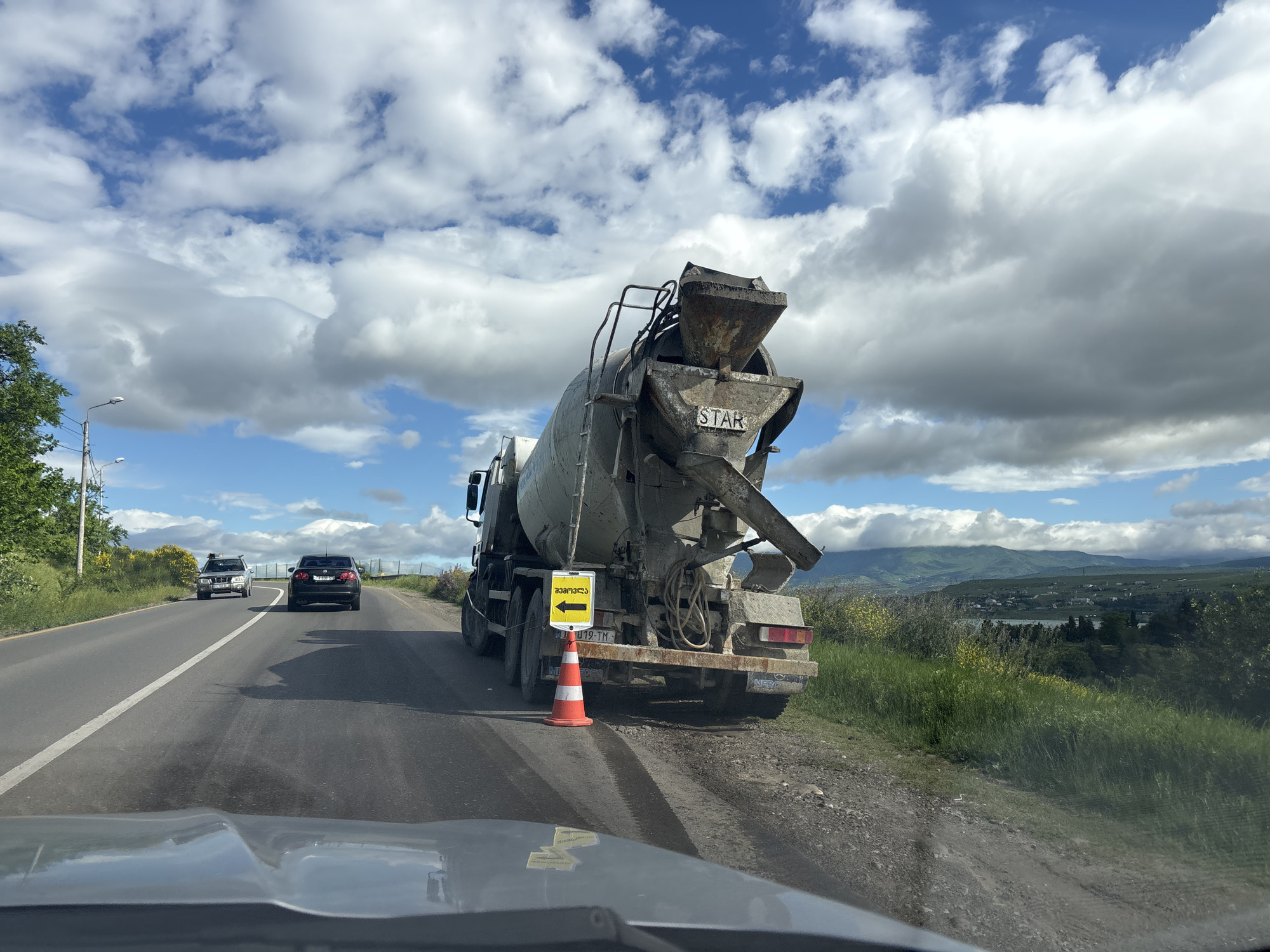 A cement truck is parked on the side of a road, partially obstructing the lane of traffic. The truck has a sign with a left-pointing arrow, indicating traffic should proceed around it. An orange traffic cone is placed behind the truck for additional visibility. Two cars are visible on the road ahead of the camera, one a dark-colored sedan and the other a silver SUV. The sky is partly cloudy. Hills and a body of water are visible in the background. Based on the truck’s license plate and the arrow in a square with Georgian writing on the sign, this photo was taken in the country of Georgia, although not necessarily in the city of Tbilisi.