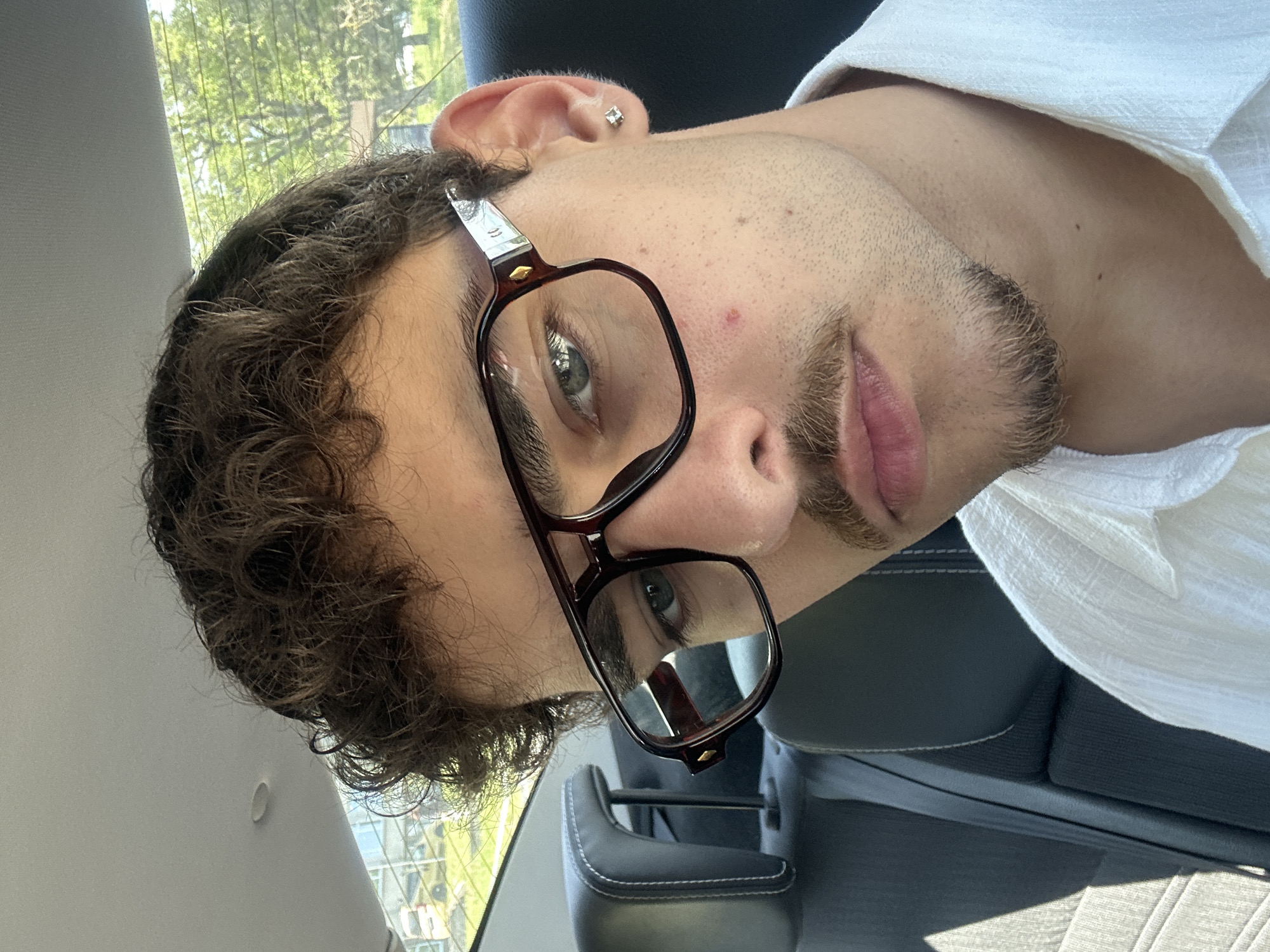 A fair-skinned individual with curly brown hair, a short beard, and glasses sits in a vehicle. The person wears a white shirt. The environment appears to be inside the vehicle. It is not possible to determine the location, including whether the image was taken in Lovech, Bulgaria.