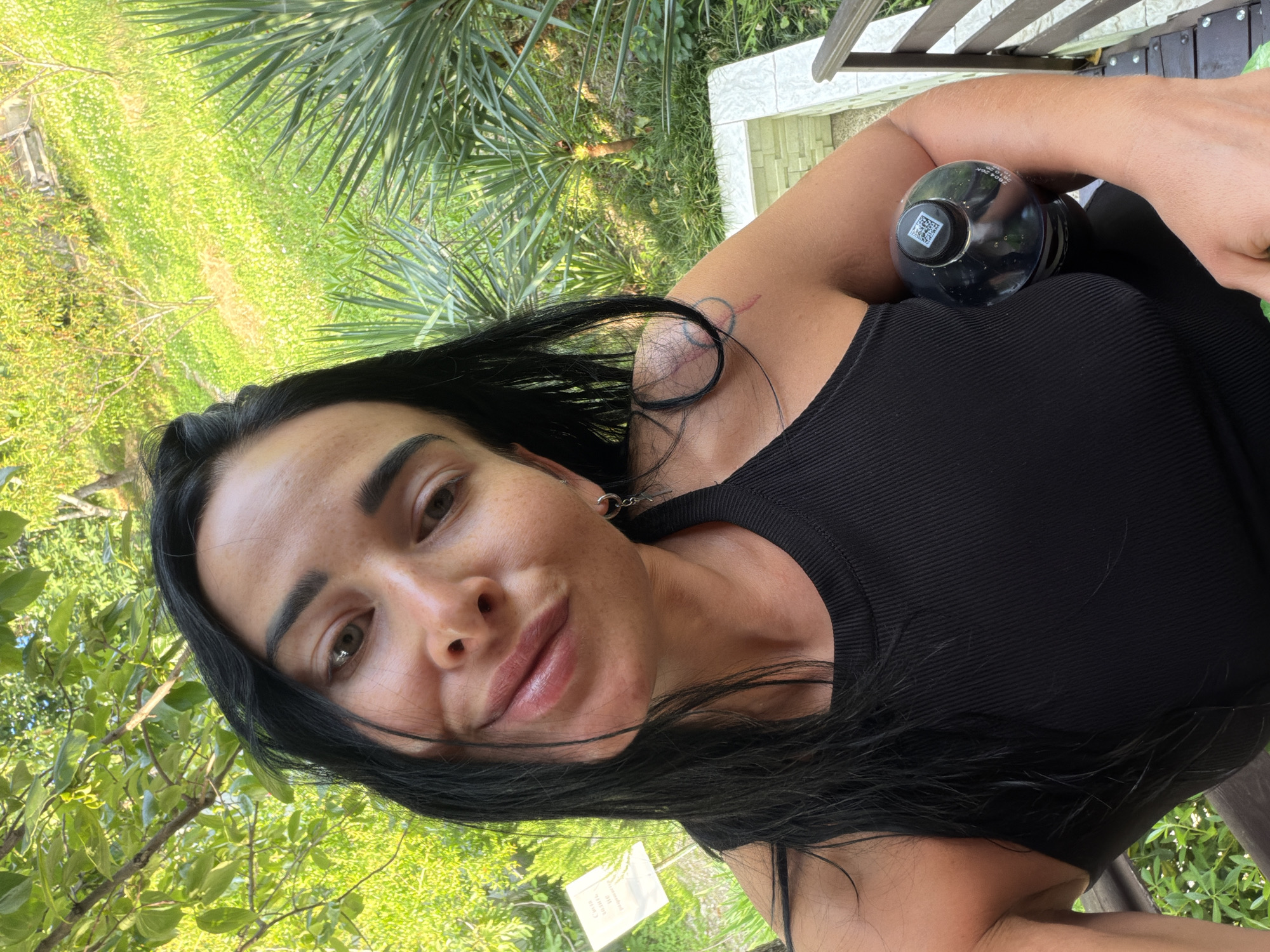 A woman with dark hair and dark tank top holds a black bottle of drink while posing for a selfie. She is outdoors with green foliage and trees in the background. The location appears to be in Sochi, Russia, given the lush vegetation and the presence of palm-like plants, common in the subtropical climate of the city. A wooden railing is visible in the lower right of the frame.