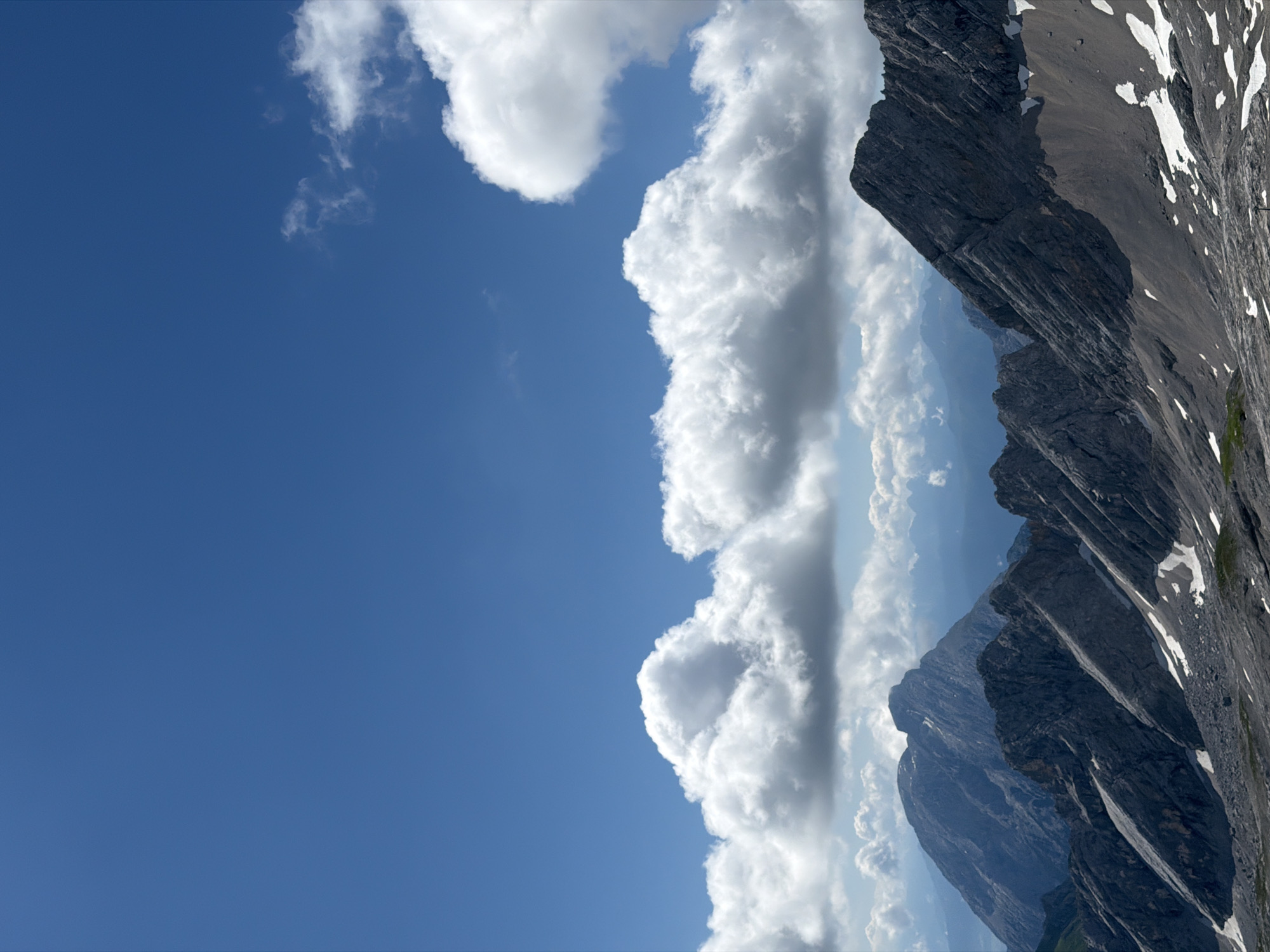 The image depicts a panoramic view of a mountainous landscape under a clear blue sky with scattered white clouds. The foreground features rugged, rocky mountain ridges with patches of snow, suggesting a high-altitude environment. In the middle ground, several more mountain peaks are visible, receding into a hazy distance. The sky occupies the upper half of the image, with the bright blue contrasting with the voluminous white clouds. This scene is characteristic of the Bavarian Alps, likely taken from a vantage point near Garmisch-Partenkirchen, Germany, a region known for its dramatic mountain scenery. There are no people or human-made objects clearly visible in the image. The overall impression is one of natural grandeur and vastness.