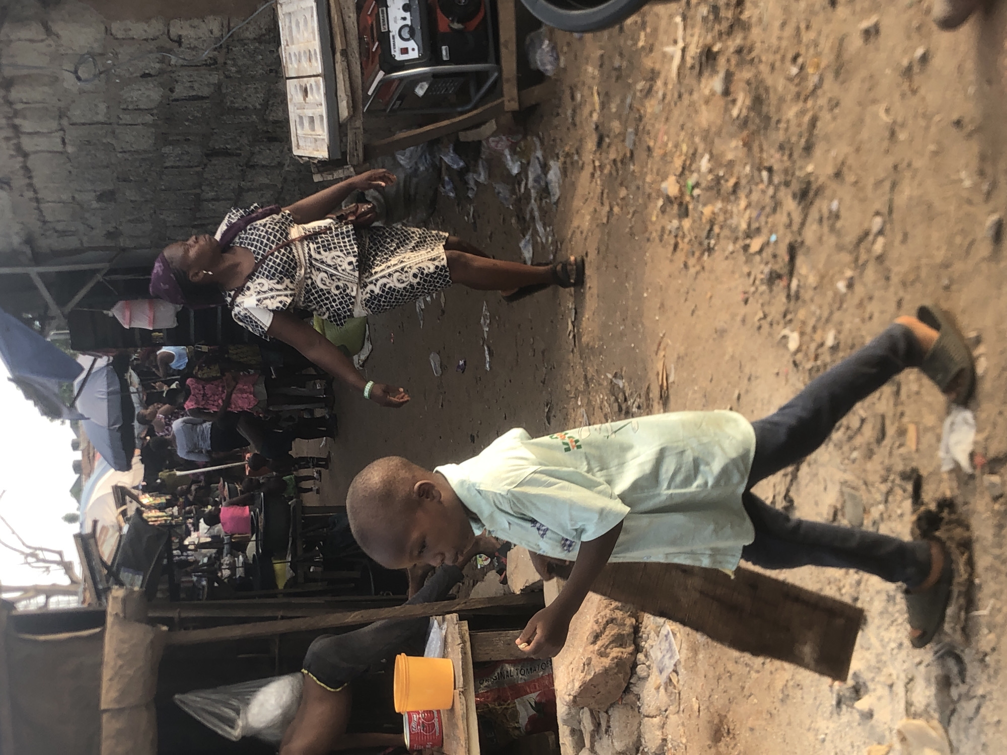 This image captures a vibrant street or market scene in Onitsha, Nigeria, characterized by an unpaved, dusty ground scattered with debris. The lighting suggests it is daytime, likely sunny.
In the foreground, a young boy with dark, short hair, wearing a light-colored shirt (possibly pale green with subtle patterns) and dark trousers, is focused on something in his hands or on the ground. He is wearing dark sandals. Near his feet are a bright yellow plastic cup and a red can. A partially visible red sack or bag reads "ORIGINAL TOMATOES."
In the mid-ground, a woman with dark skin stands with her back partially to the viewer, wearing a patterned black and white short dress and a purple headwrap. She appears to be looking towards the left side of the frame and is wearing dark sandals. Behind her, on a wooden stand, is a portable generator, predominantly black and white.
The background reveals a bustling market environment with multiple individuals, some sitting, others standing. There are simple wooden market stalls, and blue and white umbrellas provide shade over vendors or patrons. The overall impression is that of a busy, communal outdoor space. The buildings in the background appear to be constructed from stone or concrete blocks.