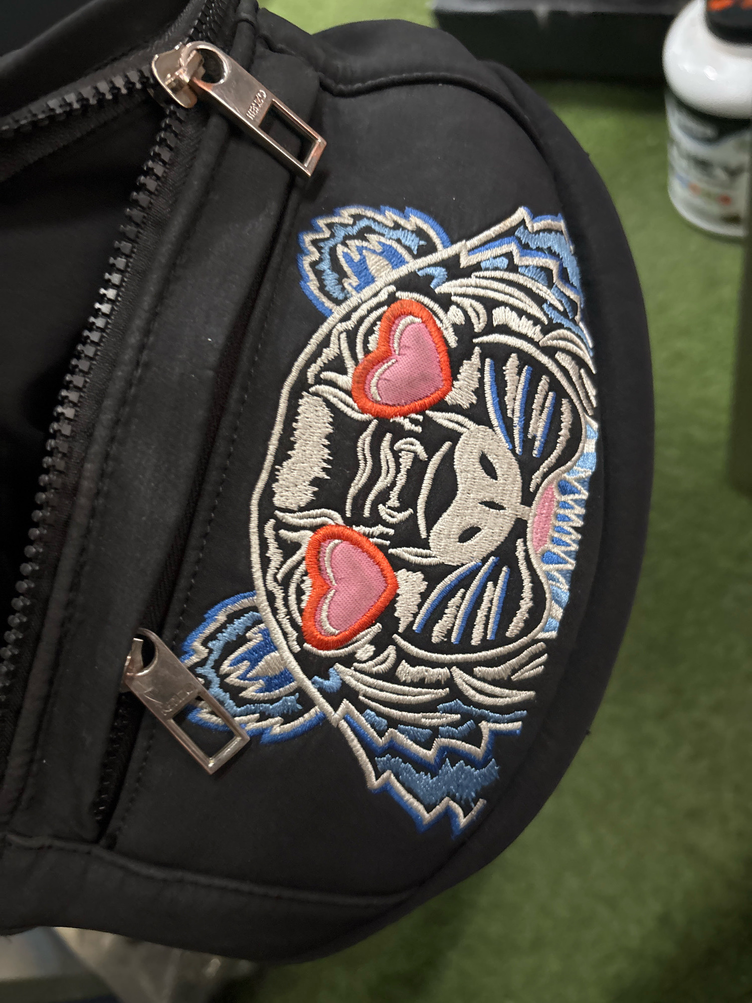 A black crossbody bag with a Kenzo tiger head embroidered on it is displayed on a green surface. The bag has silver zippers. The embroidery features the tiger's face with pink hearts for eyes, outlined in red. The background is blurred and appears to be an indoor setting with green carpeting. There is no discernible indication of the location being the city of Kepolorejo, Indonesia.