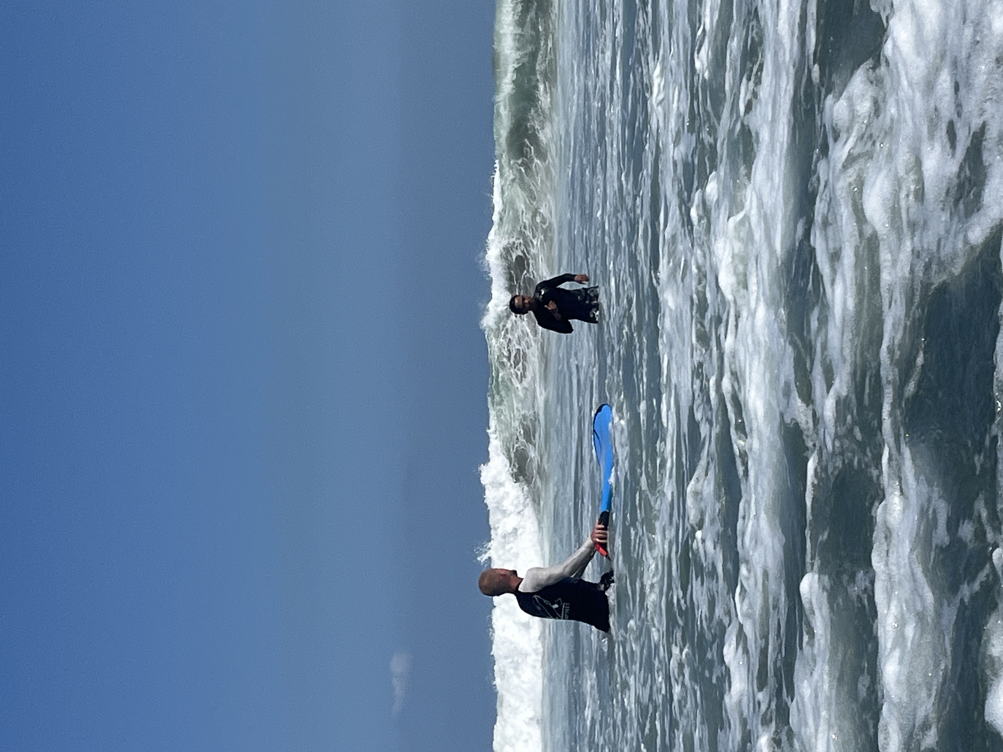 Two men are in the ocean near the coast of North Kuta, Indonesia. The man on the left, wearing a black wetsuit with white sleeves, is sitting on a blue bodyboard. The man on the right, wearing a black wetsuit, is standing in the water. Both men appear to be surfers or engaged in water sports. Behind them, waves are breaking, indicating moderate ocean conditions. The sky is clear and blue, suggesting a sunny day. The water is choppy with foam and whitecaps.