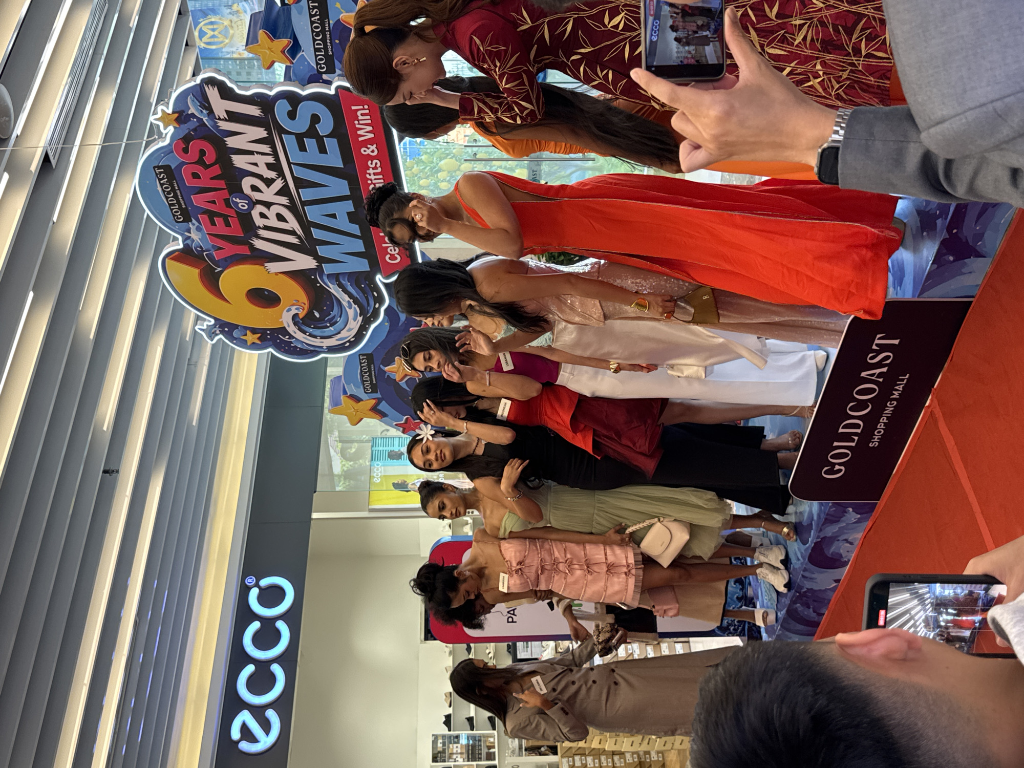 A group of women stand on a stage in what appears to be a shopping mall in Nha Trang, Vietnam. They are dressed in various formal outfits, with some holding handbags. A banner in the background advertises "6 Years of Vibrant Waves" for Goldcoast Shopping Mall. The women seem to be participating in an event or competition. In the foreground, a person's hand holds up a smartphone, recording the scene. The lighting suggests an indoor environment, likely a public space.