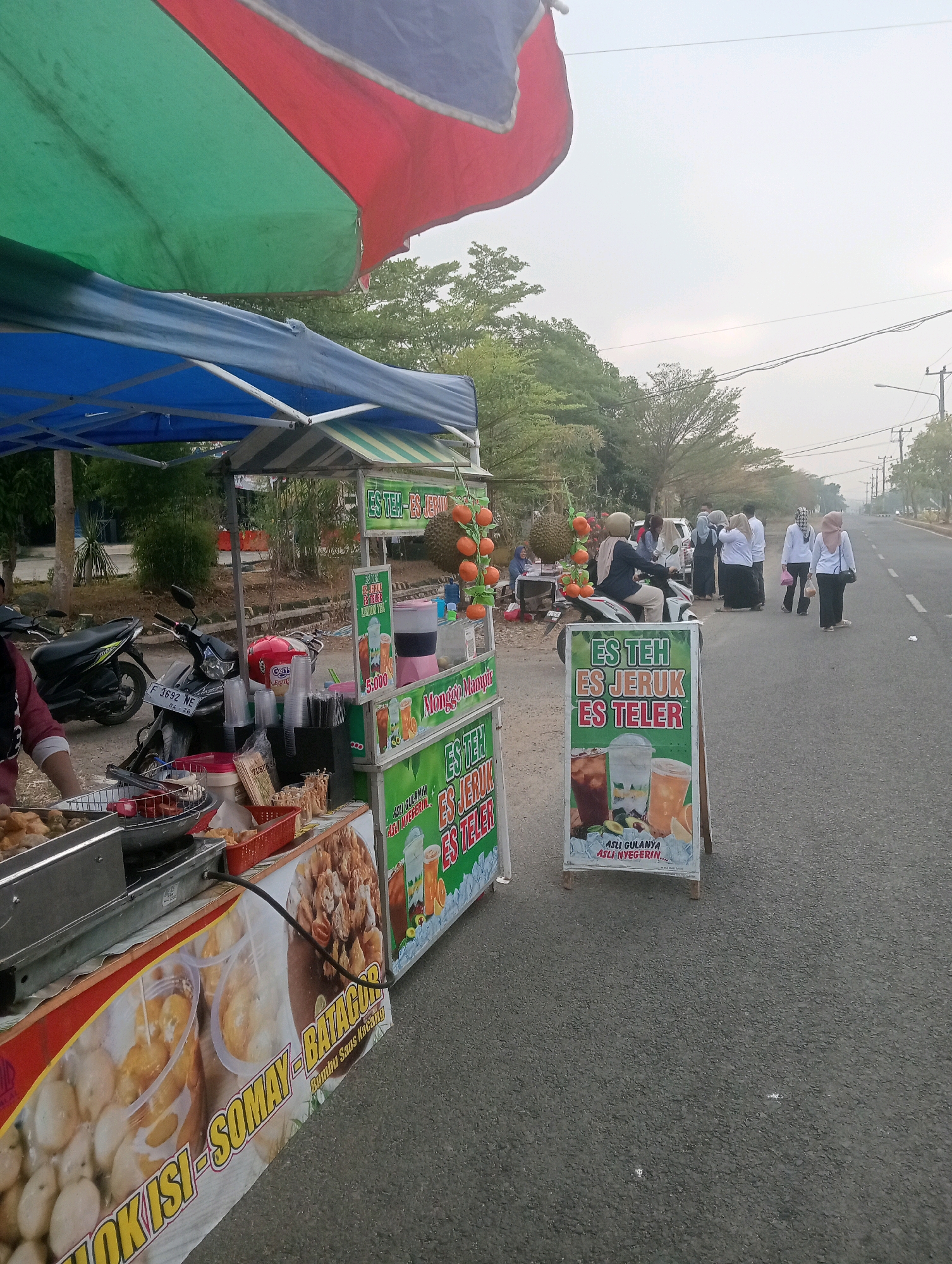 In Sidoharjo, Indonesia, an outdoor food and beverage stand is set up along a paved road under a partially visible red, green, and blue striped umbrella. The stand features multiple display boards advertising "ES TEH" (Iced Tea), "ES JERUK" (Iced Orange), and "ES TELUR" (Egg Iced Drink). One board includes images of the drinks and text indicating "ASLI GULANYA ASLI NYEGERIN" (Genuine sweetness, genuinely refreshing). The stand also displays food items such as fried snacks and what appears to be batagor and somay. Several motorcycles are parked behind the stand. In the background, a group of people, mostly women wearing headscarves and some in white shirts, are gathered near a motorcycle. The environment is characterized by trees and foliage behind the stand, and utility poles and streetlights line the road, suggesting an urban or semi-urban setting. The sky is overcast, indicating a cloudy or hazy day.