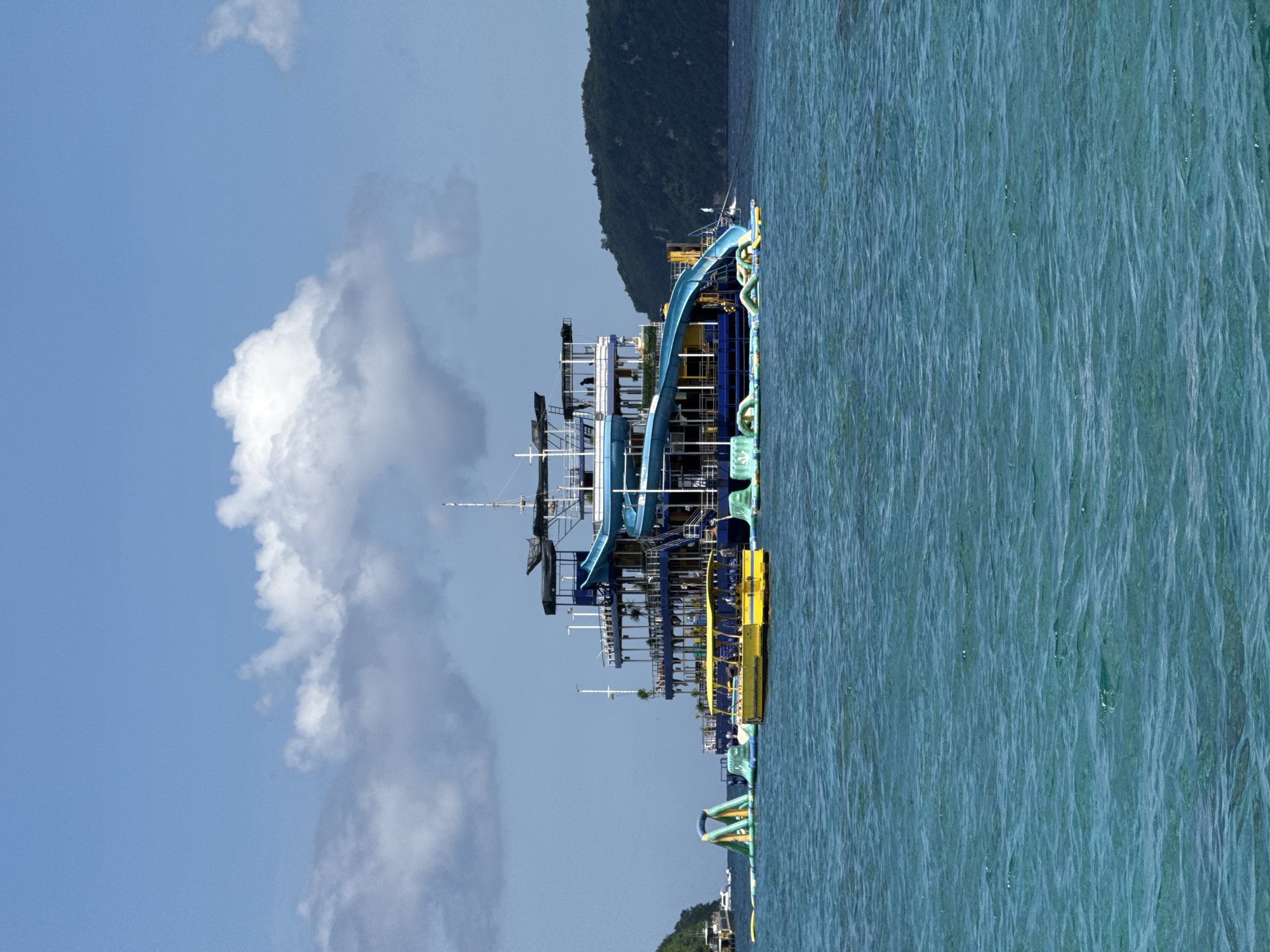 The image depicts a multi-level boat floating on the water near Nusa Penida, Indonesia. The boat features a blue slide and yellow platforms. Behind the boat, a verdant, hilly landscape stretches into the distance. To the left of the boat, the sky is a pale blue with large white clouds. The water is a vibrant turquoise, reflecting sunlight and showing surface ripples.