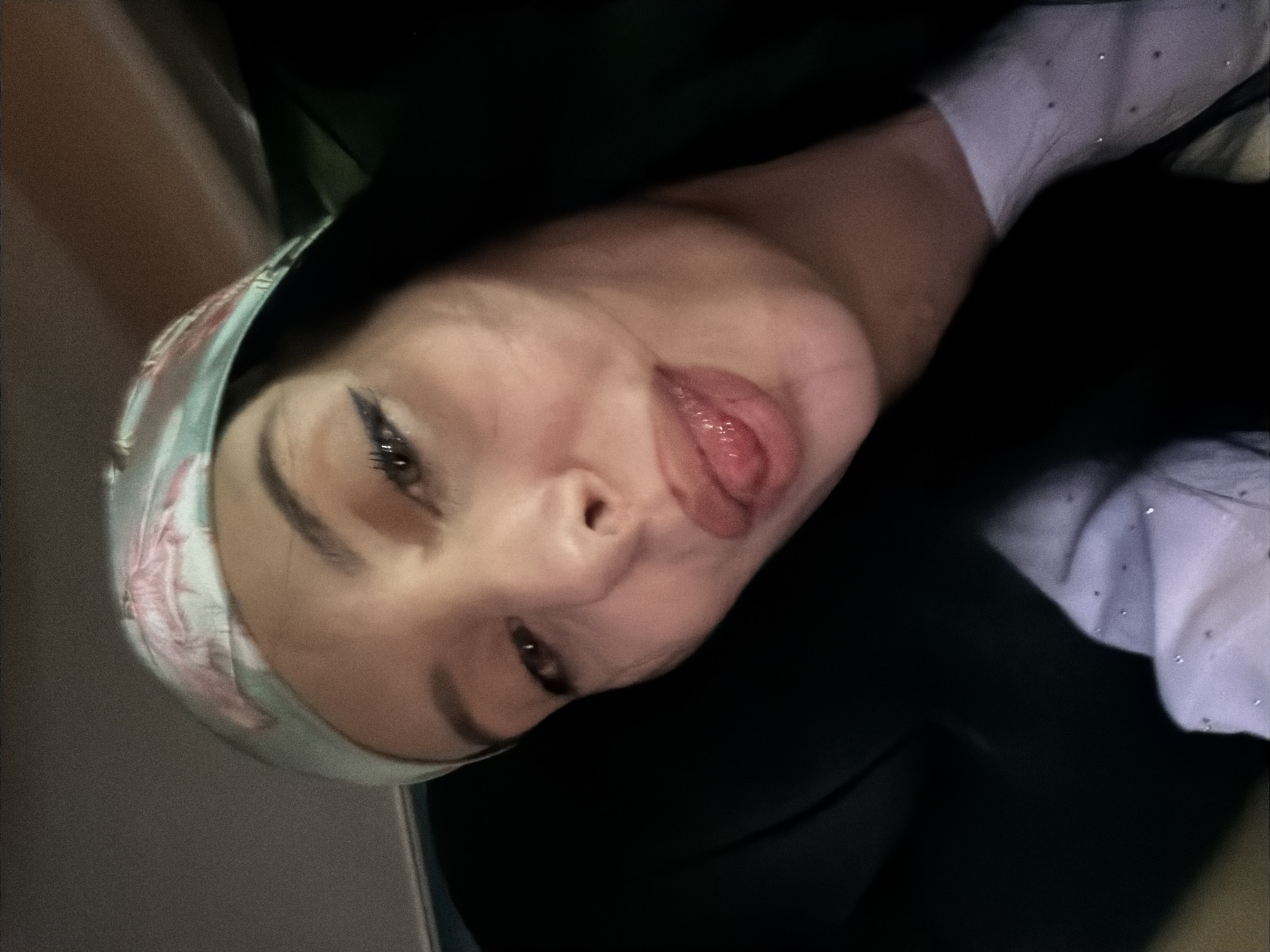A close-up, selfie-style photograph depicts a young woman from the chest up, indoors, with her head tilted and her tongue playfully extended. She is wearing a light blue headscarf adorned with a floral pattern, possibly depicting pink flamingos. Her makeup includes winged eyeliner and lip gloss. She has long, dark hair and is wearing a light purple shirt with small, shimmering embellishments. The background is dark and out of focus, with a hint of a dark chair or seat. There is no discernible information in the image to suggest a location in Tbilisi, Georgia.