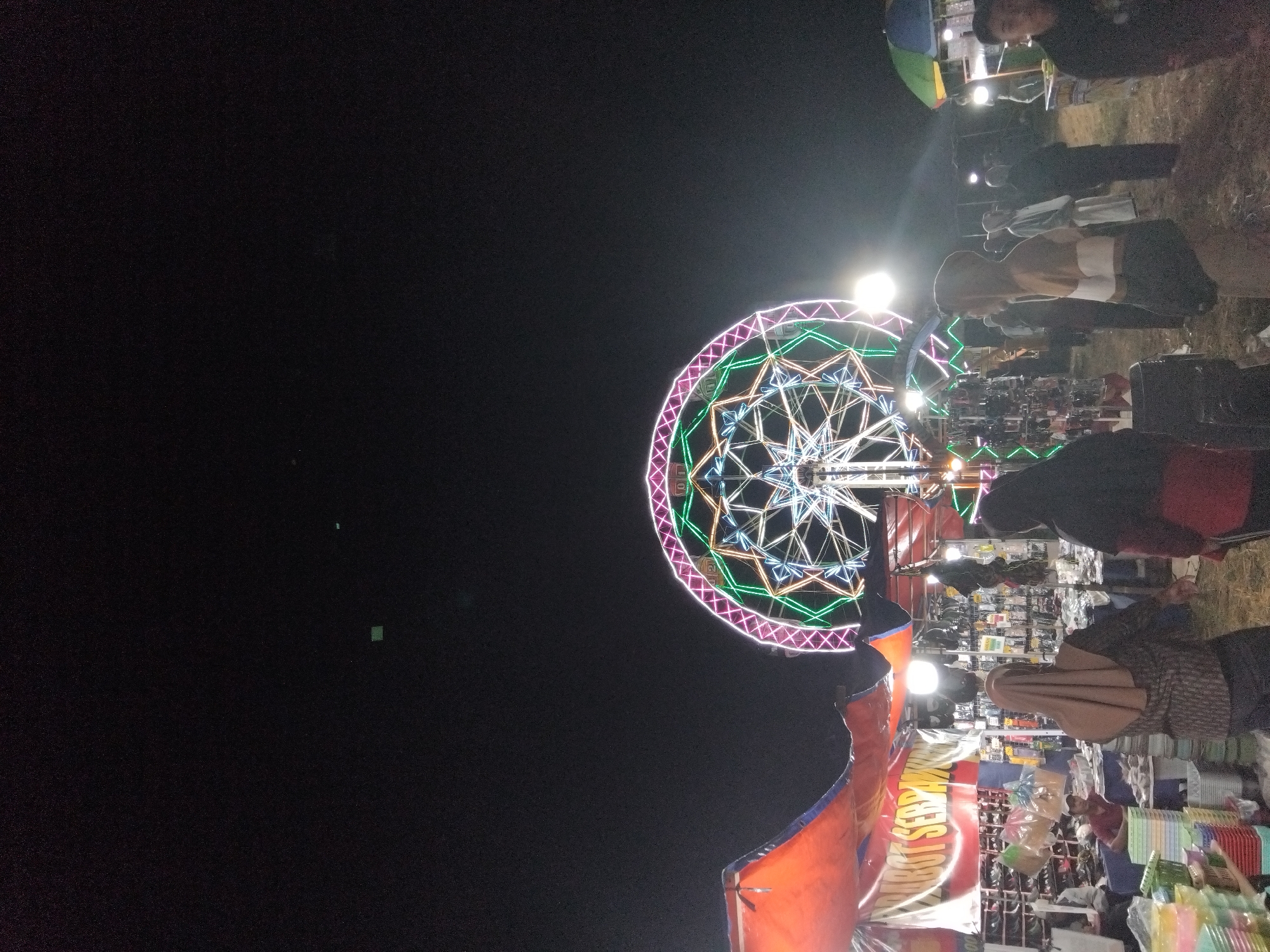 The image depicts a night scene at a fair or market in Sambirata, Indonesia. A large ferris wheel, illuminated with colorful neon lights in pink, green, and white, dominates the background. In the foreground, there are various stalls. One stall on the left features an orange awning with a banner that appears to read "NEBOT SERPANT." The stall is laden with merchandise, including stacks of colorful plastic containers and shelves filled with various items. Several people are visible, some browsing the stalls, and others walking through the fair. The ground appears to be a dirt or sandy surface. The overall atmosphere suggests a lively community event taking place after dark.