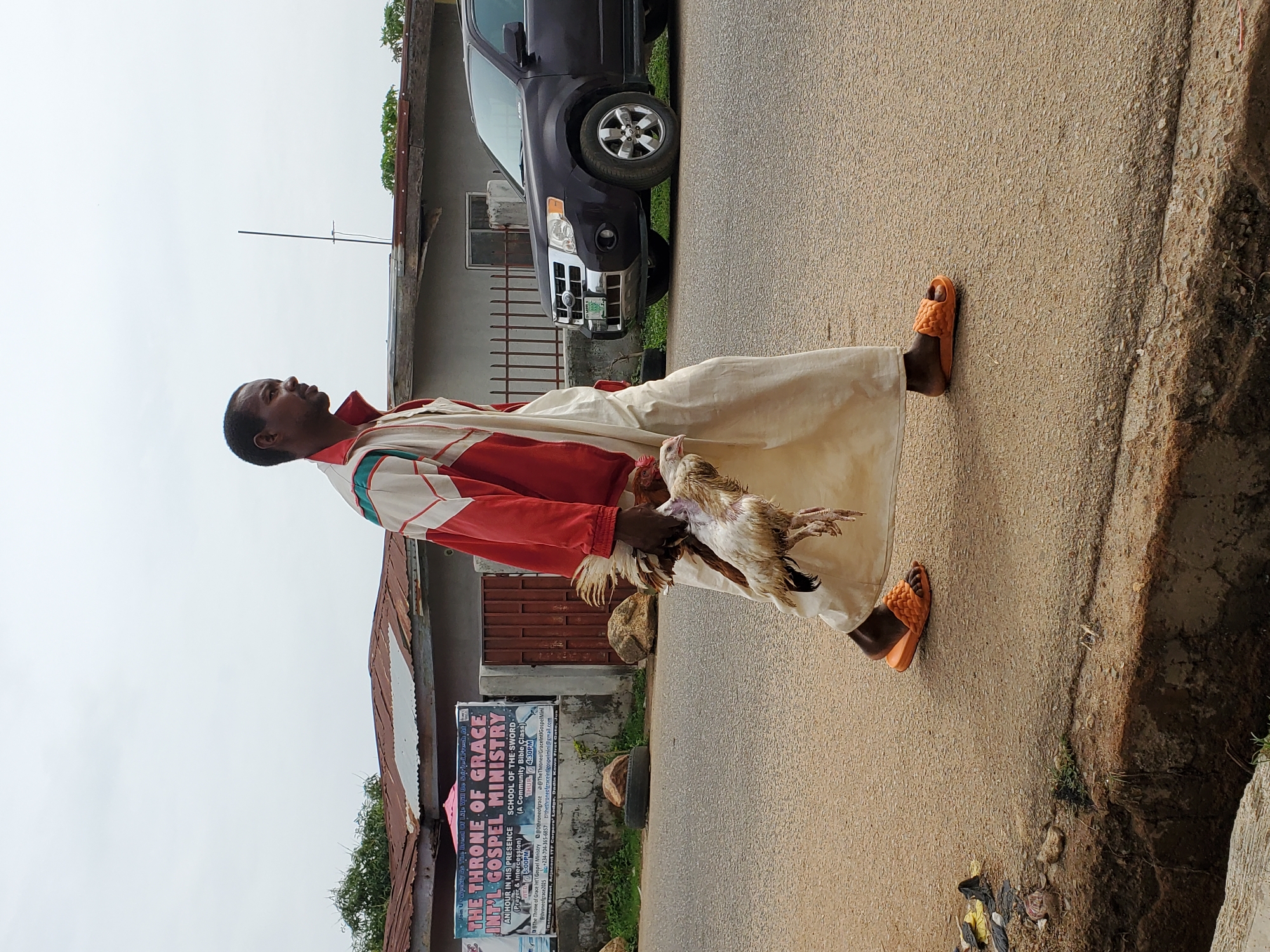 A man walks down a paved street carrying a dead chicken. The man is wearing orange sandals, loose off-white pants, a red and white jacket, and looks upwards. To the right of the man is a raised, uneven ground with plants growing. To the left of the man is a sign indicating “The Throne of Grace Int’l Gospel Ministry." The sign also includes the name "Dadin Kowa" which locates the area to Jos, Nigeria. In the background, a dark-colored SUV is parked on the side of the road in front of a building with a corrugated metal roof.