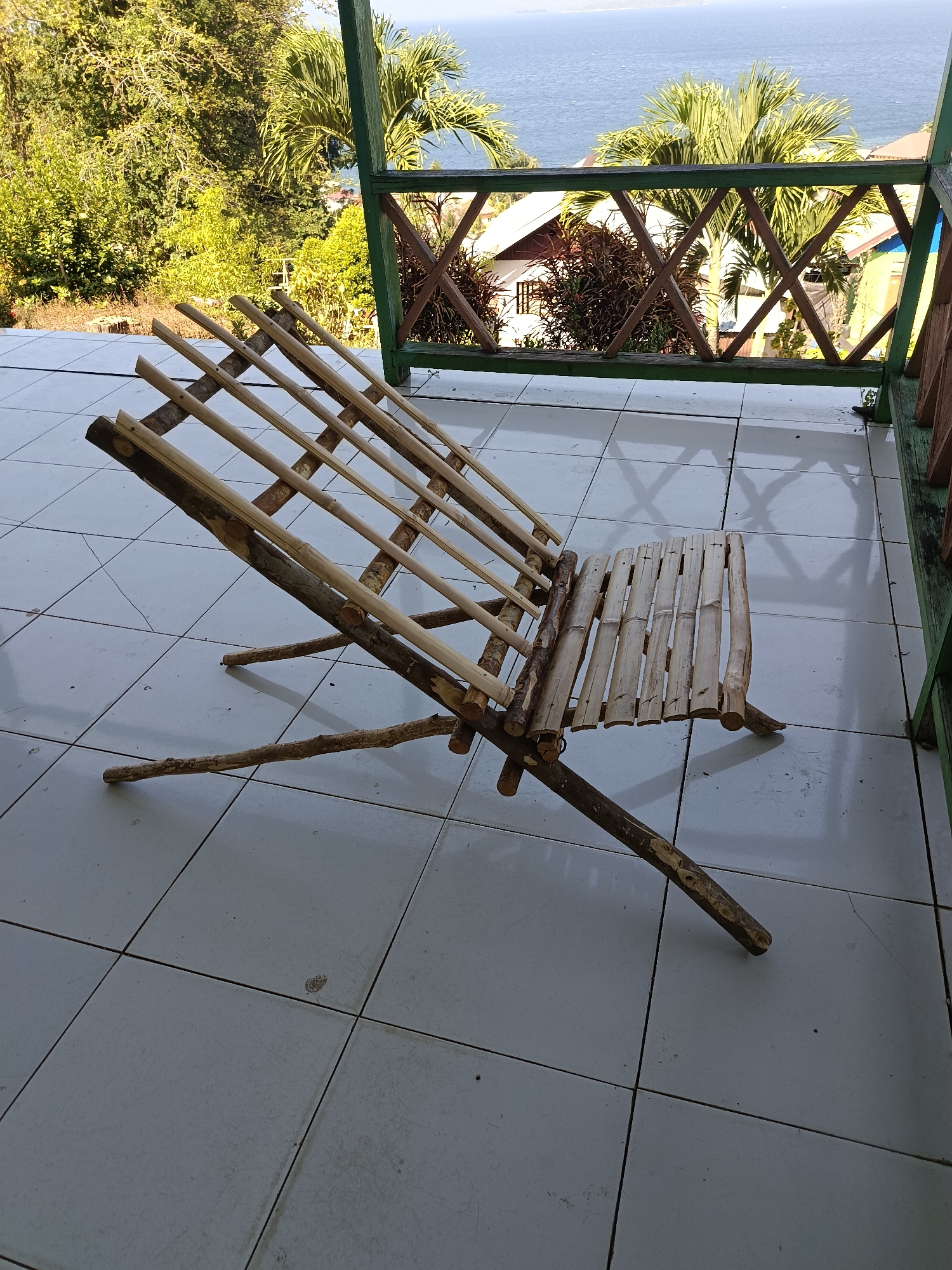 A handmade wooden lounge chair made of bamboo and branches is situated on a white tiled patio overlooking a body of water. The chair is angled, with its backrest made of several parallel bamboo poles and its seat constructed from a series of shorter, wider bamboo pieces. The chair's legs and supports are made from thicker branches. The patio tiles are laid out in a grid pattern and are somewhat weathered.
In the background, beyond the patio, a green wooden railing with a diamond-patterned infill separates the patio from the view. Beyond the railing, lush green trees and palm trees are visible. A glimpse of houses with white walls and terracotta roofs can be seen nestled among the foliage. The expansive blue water of the sea stretches to the horizon under a clear sky. The scene suggests a tranquil, tropical setting, likely in Mansalean, Indonesia, where such natural materials and elevated positions overlooking the ocean are common. The lighting indicates it is daytime, with the sun casting distinct shadows on the tiled surface.
