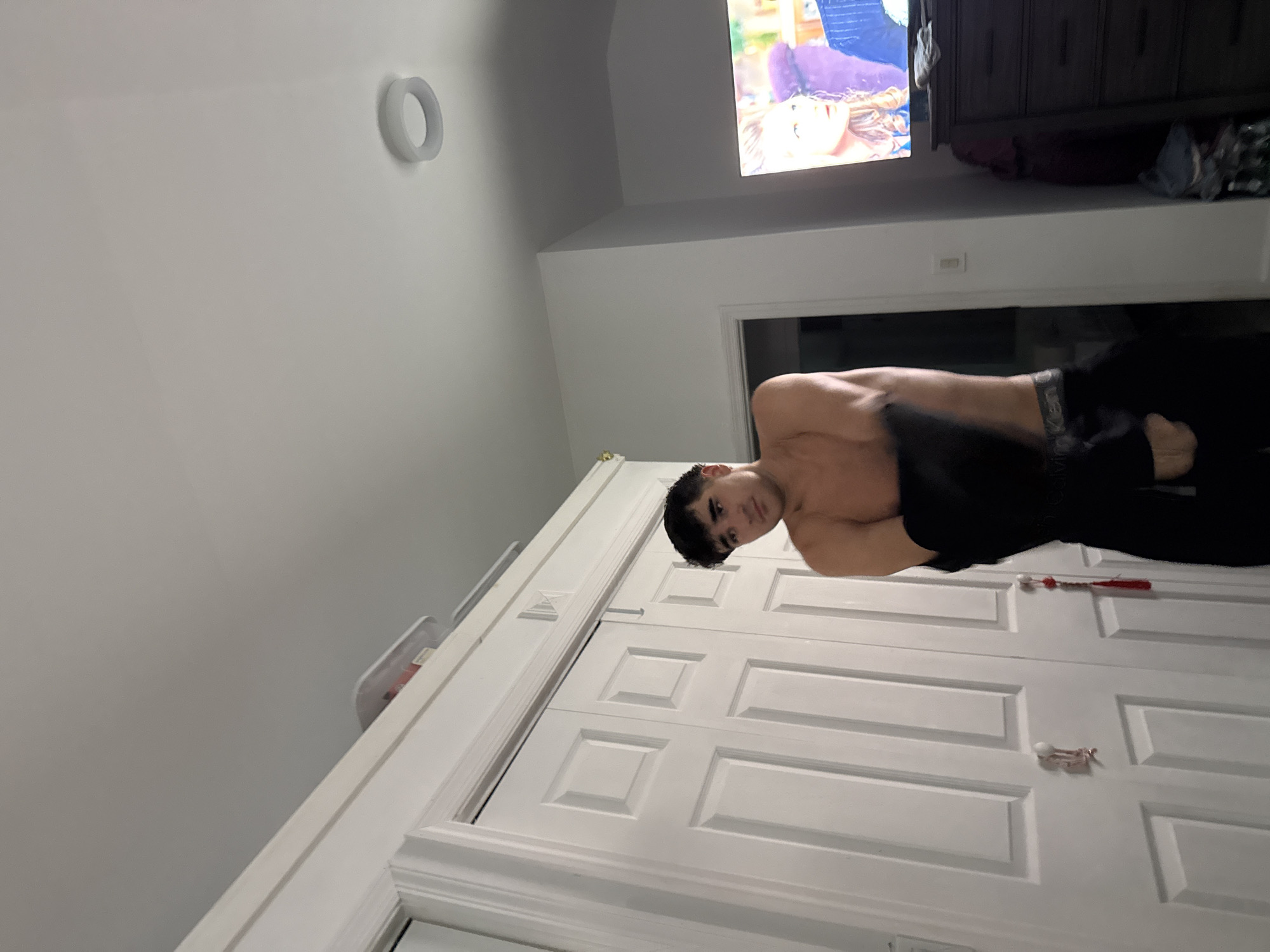 A young man is partially undressed in a room with white double doors in the foreground and a dresser with a television on top in the background. The man is facing left, pulling a dark garment over his head. He is wearing black underwear with "Calvin Klein" visible. The room appears to be a bedroom, with a light fixture on the ceiling. A doorway is visible to the right of the man. The television screen shows an image of two people. A white container with red accents is on top of the double doors. There is a red tassel hanging from one of the door handles. The location is Bellmore, United States of America.