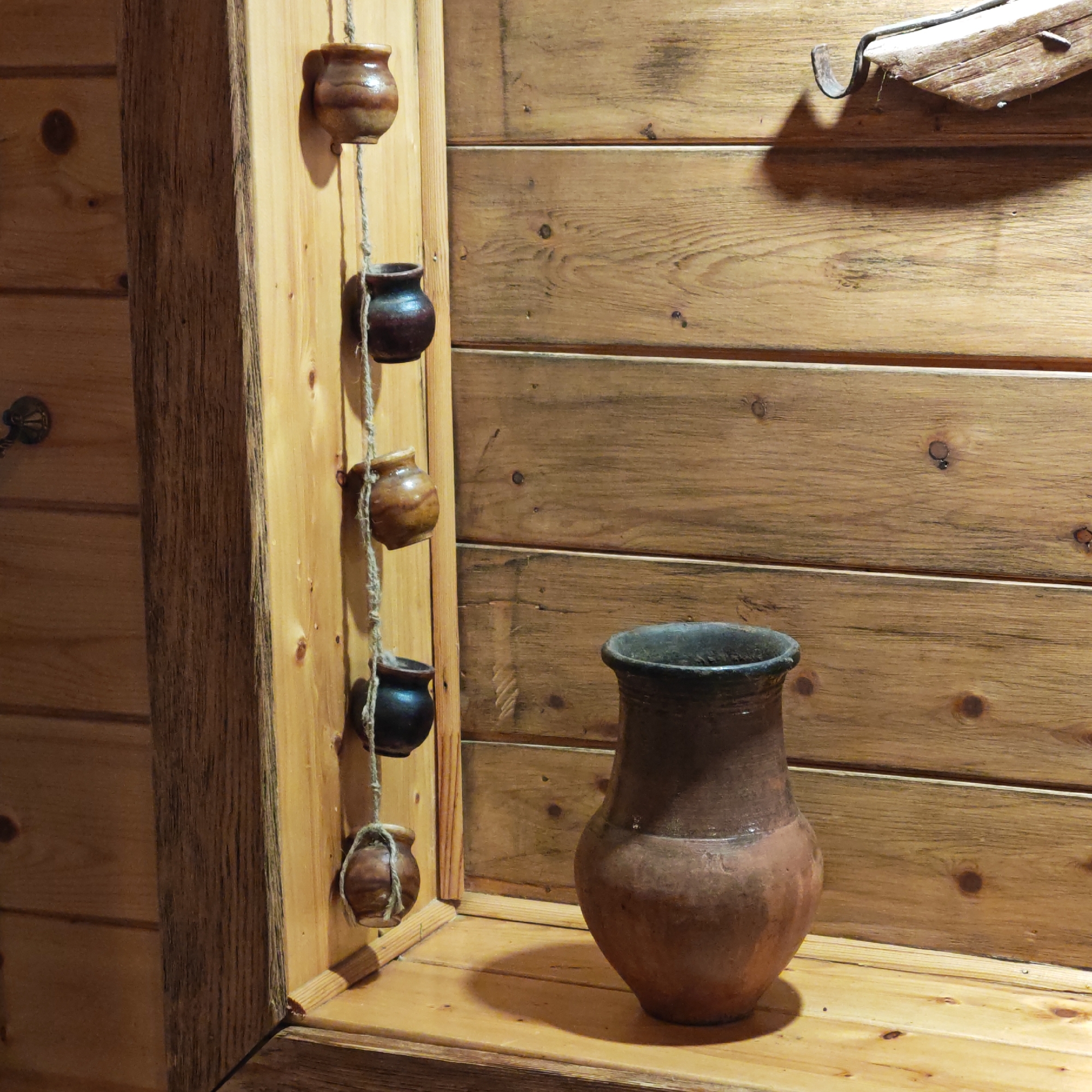 The scene is a rustic interior, likely in the city of Rostov, Russia, given the provided context. The walls are clad in horizontal wooden planks of a light brown hue. In the lower right of the frame, a large, earthenware jug, brown with a darker rim, sits on a wooden surface. To the left, hanging from a wooden beam, are five small ceramic pots suspended by twine. The pots vary in color from light brown to dark brown. The beam itself is integrated into the wooden wall, contributing to the overall rustic aesthetic. An indistinct, dark, wooden object is partially visible in the upper right corner. The lighting is ambient and warm, casting soft shadows.