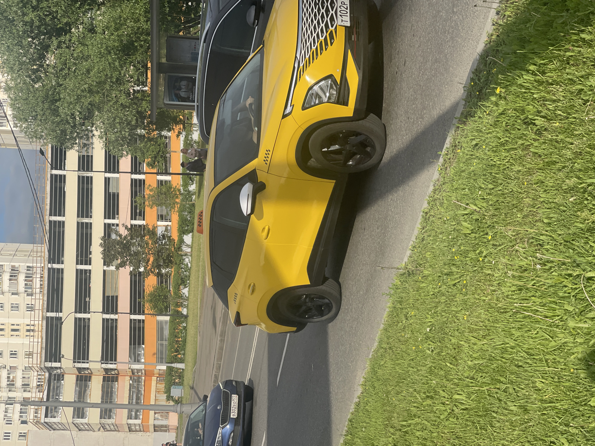 This image, originally rotated 90 degrees counter-clockwise, depicts a bright daytime urban street scene in Moscow, Russia.
In the foreground, a vibrant yellow taxi SUV, possibly a Moskvich 3, is prominently parked on the right side of a road or parking area. The vehicle features a distinctive black-and-white patterned grille, black multi-spoke wheels, and a white side mirror. A black and white checkered taxi stripe is visible on its front fender. The Russian license plate on the front reads "T102P 777". The driver is partially visible through the windshield.
To the left of the yellow taxi, a dark blue car, likely a Skoda model given faint emblem hints, is parked further down the street. Its Russian license plate, "B302EO 977", confirms the Moscow region. Another dark vehicle is partially visible parked in front of the yellow taxi.
The background features several multi-story buildings, typical of urban residential or commercial architecture. They display various pastel colors, including beige, light pink, and orange, and have numerous rectangular windows. One of the buildings appears to have scaffolding erected on its side. Lush green trees are scattered throughout the background, adding foliage to the urban landscape.
Near the yellow taxi and the buildings, a bus stop or public transport shelter is visible, with an advertisement poster that includes some unreadable Russian text, possibly mentioning "в Москве" (in Moscow). Two people are standing near this shelter. Further in the background, near the blue car, a few other pedestrians can be seen.
The scene is well-lit by bright sunlight, suggesting late morning or afternoon. The weather appears clear and sunny, casting distinct shadows on the asphalt and pavement. A strip of green grass, dotted with small yellow flowers, runs along the edge of the street on the far right.