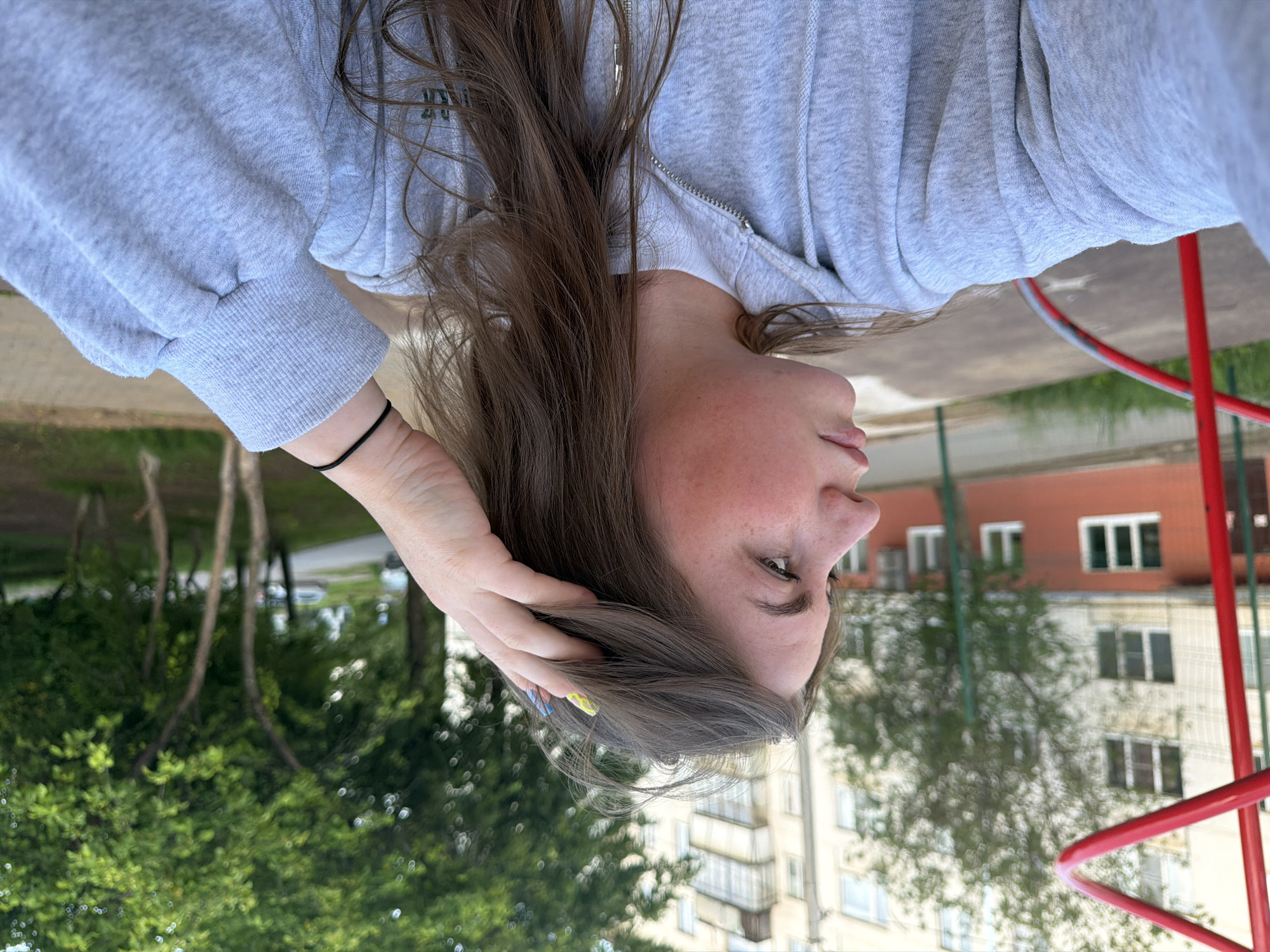 A medium-close-up shot, oriented vertically, captures a young individual from a low angle, facing slightly away from the camera. The subject has long, flowing brown hair, some of which is held by her left hand near her ear. Her skin shows freckles, and her expression is contemplative, with her gaze directed to the side. She wears a light grey hooded sweatshirt with a zipper front, and a black elastic hair tie is visible on her left wrist. Her thumb has a bright yellow nail polish, and another nail appears to have a blue or green polish.
The background reveals an outdoor setting, likely a residential area in Chaikovsky, Russia, given the context. Behind and to the side of the subject, tall apartment buildings with multiple windows are visible. Some buildings are red-brick, while others are lighter-colored. Lush green trees and foliage dominate the midground and foreground, suggesting a park or courtyard environment. Parts of red metal structures, possibly from a playground or sports equipment, are seen on the right side of the frame, providing a contrasting pop of color against the greenery and buildings. The lighting appears soft and diffused, indicating daytime, possibly overcast conditions. No visible text is present in the image.