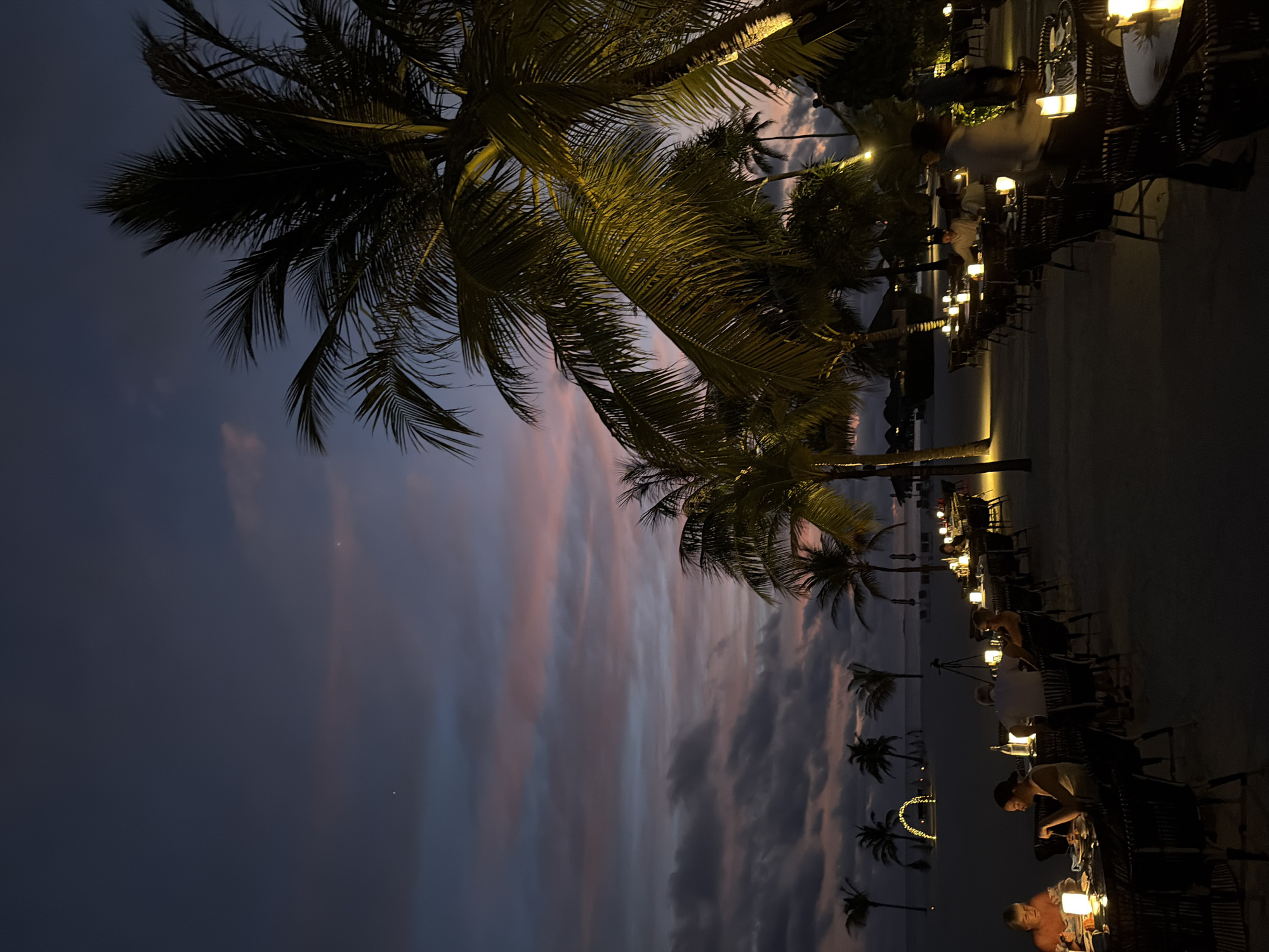 The image depicts an outdoor dining scene at dusk on a beach in Halaveli, Maldives. Several tables are set up on the sand, illuminated by candlelight and soft outdoor lighting. Palm trees are visible throughout the scene, with the sky above displaying hues of purple, pink, and dark blue as the sun sets. Silhouettes of palm trees are also present in the background, along with some resort structures and a distant archway decorated with lights.
People are seated at some of the tables, engaged in dining and conversation. The overall atmosphere is serene and romantic, characteristic of a tropical evening resort setting. The lighting, the tropical foliage, and the presence of dining tables on the beach strongly suggest a resort restaurant operating in the evening.