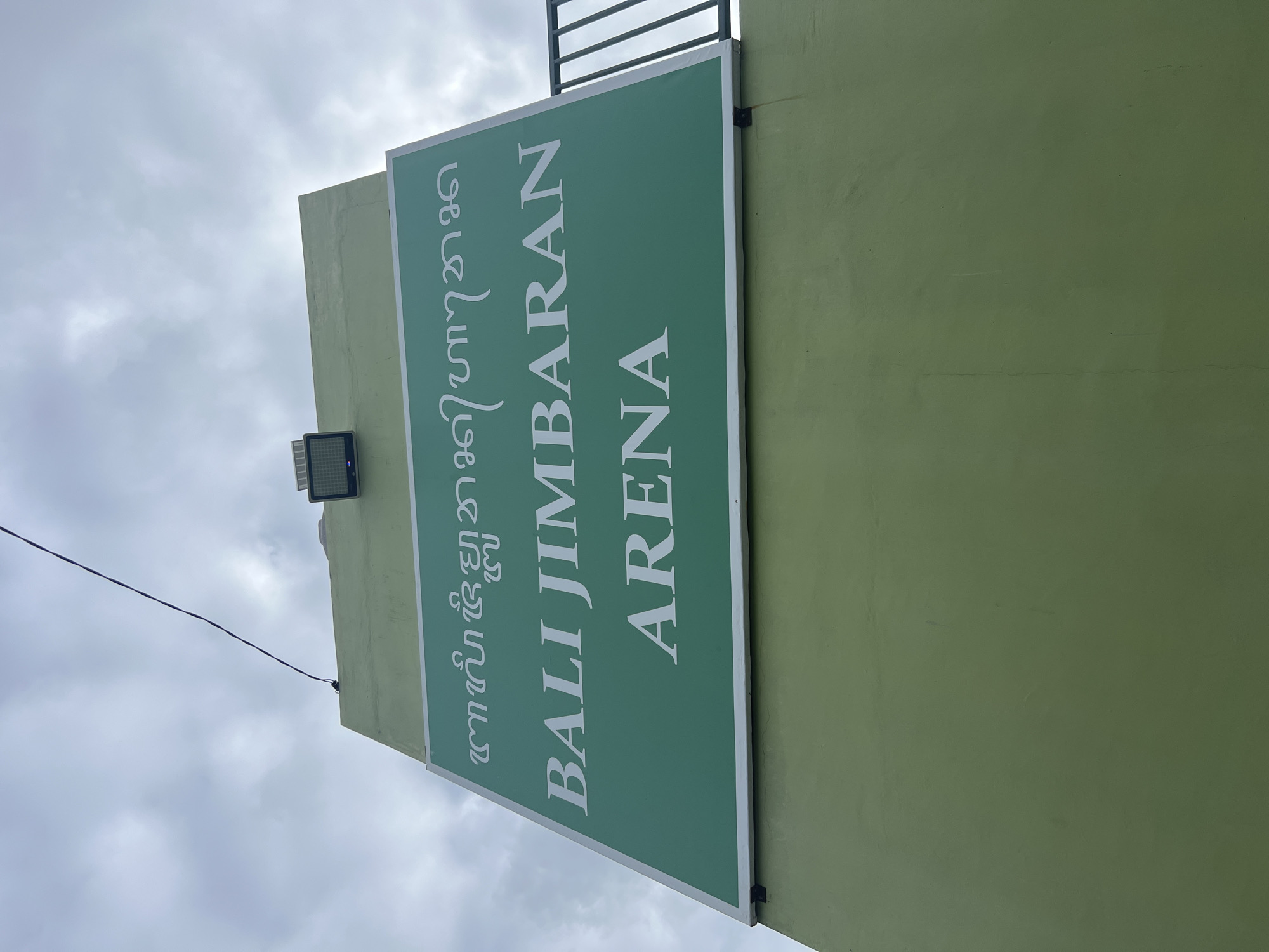 A green sign with white lettering is mounted on a light green wall, angled upwards towards a cloudy sky. The sign reads "BALI JIMBARAN ARENA" in larger, sans-serif font on the bottom two lines. Above this, in a smaller, stylized script, are what appear to be Balinese characters. A spotlight is mounted on the top edge of the wall structure. The background is a dull, overcast sky with some visible clouds. The sign is fixed to the wall with visible metal brackets at the bottom. The scene is captured from a low angle, looking up at the sign and sky. The location is Kuta Selatan, Indonesia.