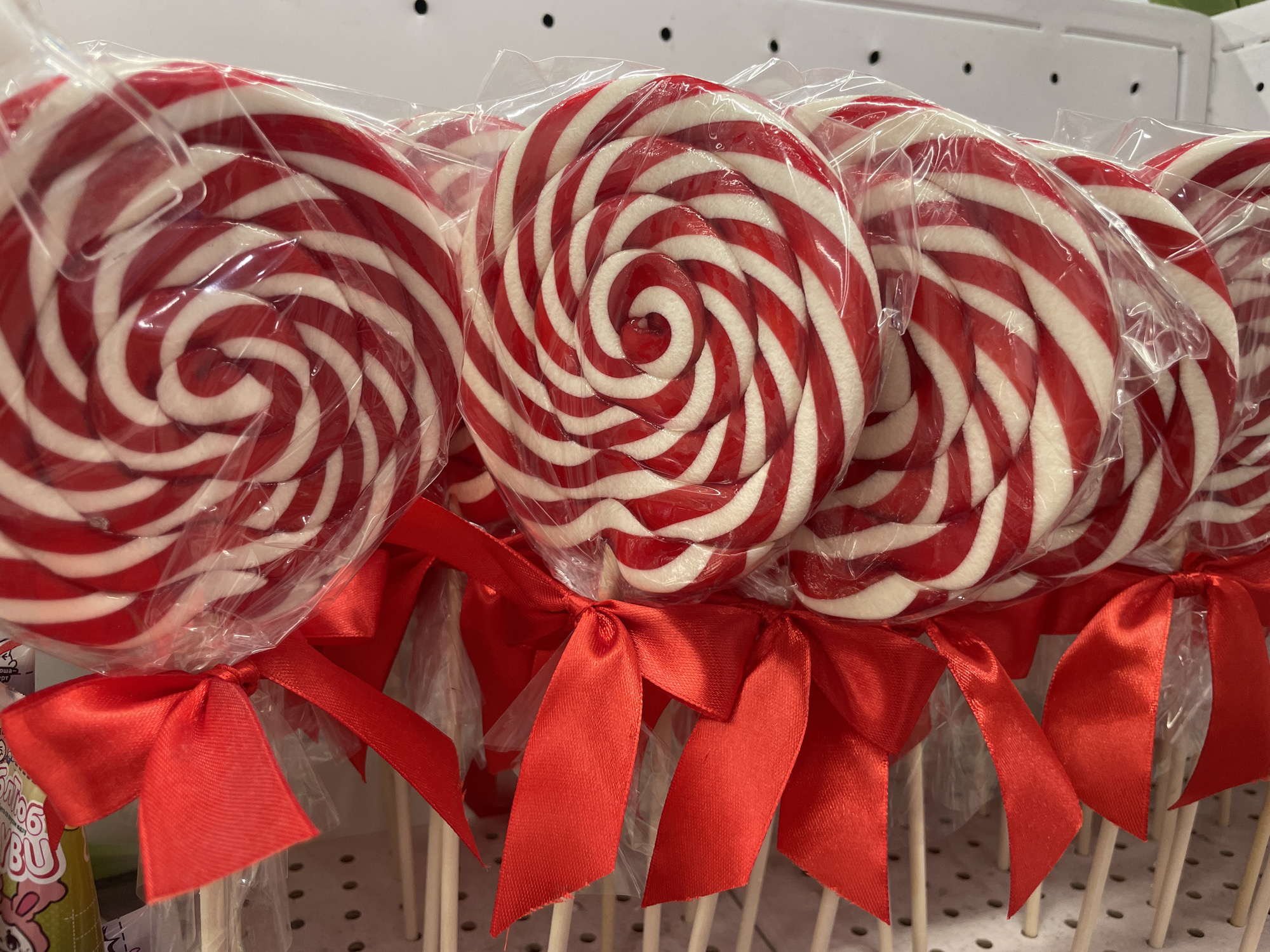 A row of large, individually wrapped, red and white swirled lollipops is displayed. Each lollipop is on a wooden stick and tied with a red satin ribbon bow. The lollipops are arranged in the foreground, with their plastic wrappers reflecting the light. In the background, shelves with small, blurred objects and some visible text can be seen. The overall scene suggests a retail or market setting, possibly a confectionery store. The prominent red and white colors of the lollipops, along with the ribbons, evoke a festive or holiday atmosphere. No people are visible in the image. There is some Cyrillic text visible on a green object to the left of the lollipops, but it is too blurry to read. The lighting suggests an indoor environment.