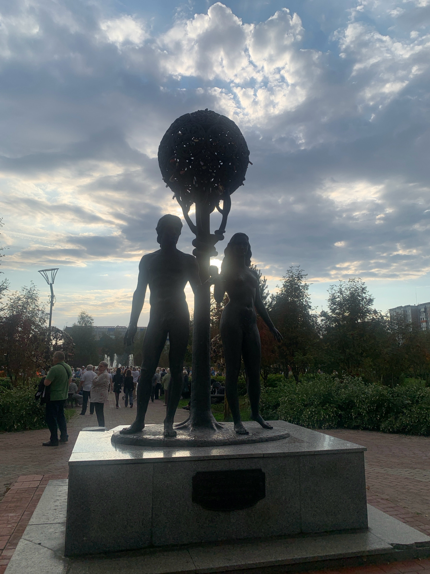 The image shows a bronze sculpture of a male and female figure standing on a pedestal. The figures appear to be Adam and Eve. Above them is a globe-like structure made of metal. The sculpture is located in Prokopyevsk, Russia, in a park. In the background, there are trees, buildings, a lamppost, and people walking. The sky is cloudy with patches of sunlight.