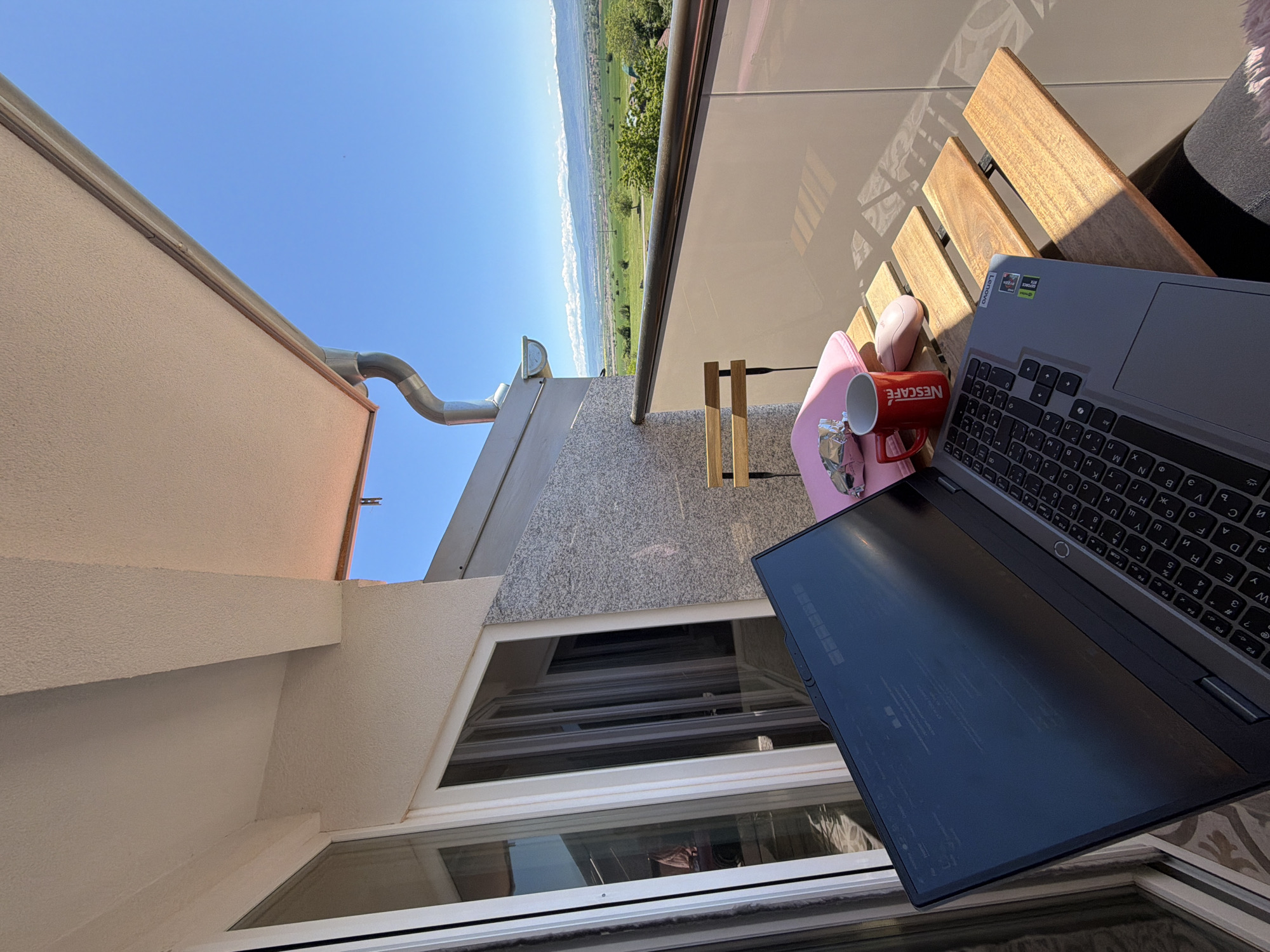 The image captures a scene on a balcony in Buhovo, Bulgaria, with a partial view of the landscape. A laptop, a red mug with the brand "NESCAFÉ", and a pink object are placed on a wooden chair. A building structure is on the left, with a metal pipe extending from it. A scenic view of fields and mountains is visible in the background under a clear sky. The overall setting suggests an outdoor working environment on a sunny day.