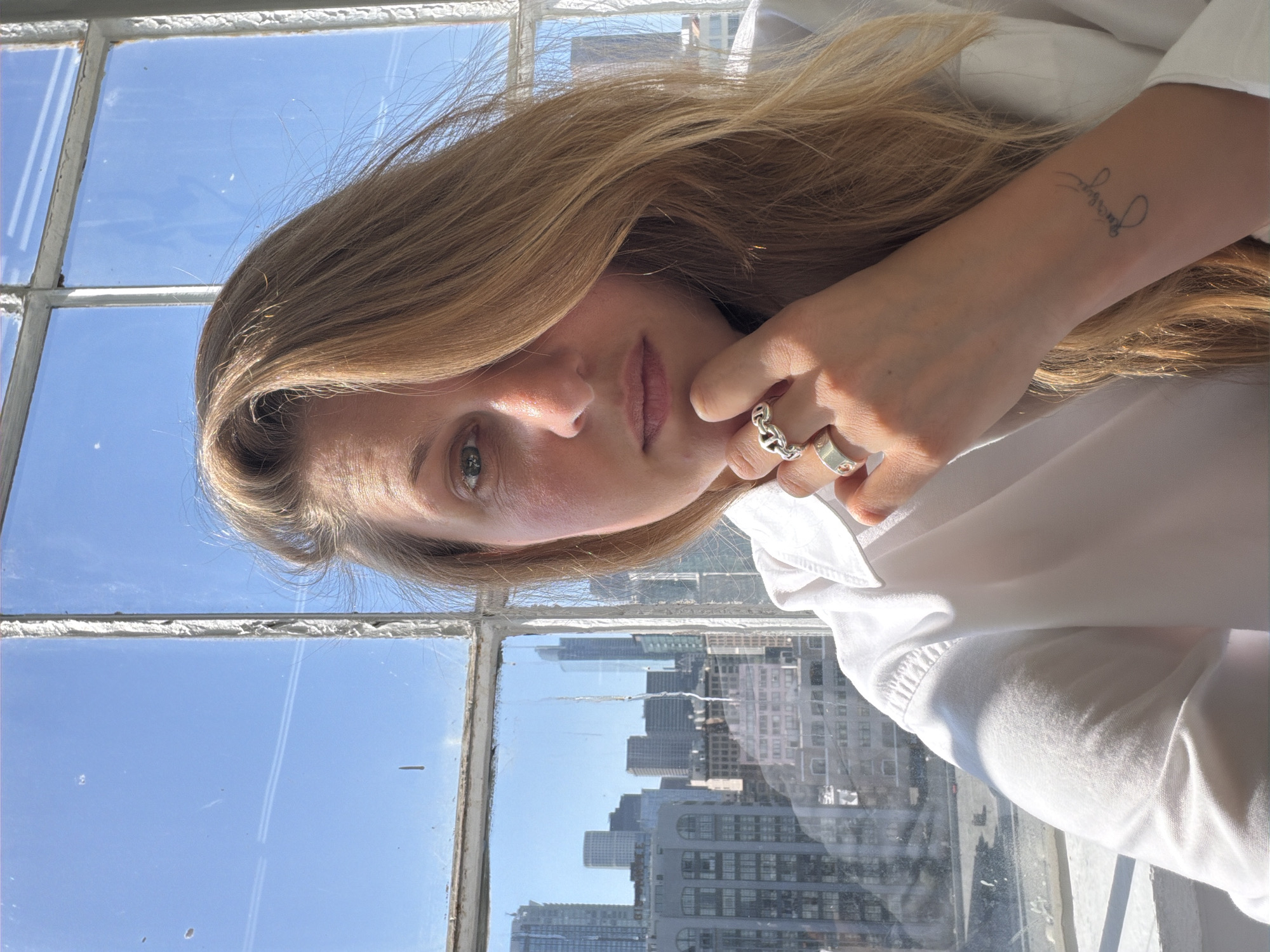 A young woman with light brown hair is positioned in front of a window that looks out onto a cityscape. The window has multiple panes, with a clear blue sky visible through some and the tops of buildings visible through others. The buildings are densely packed and appear to be in a city. The woman is wearing a white shirt. Her left hand is near her face and on her wrist is a tattoo. Her face is angled, and she is looking directly at the viewer. She wears two rings on her fingers. The scene is well-lit, suggesting it is daytime. The style of the buildings and the clear sky suggests that this could be Los Angeles, United States of America.