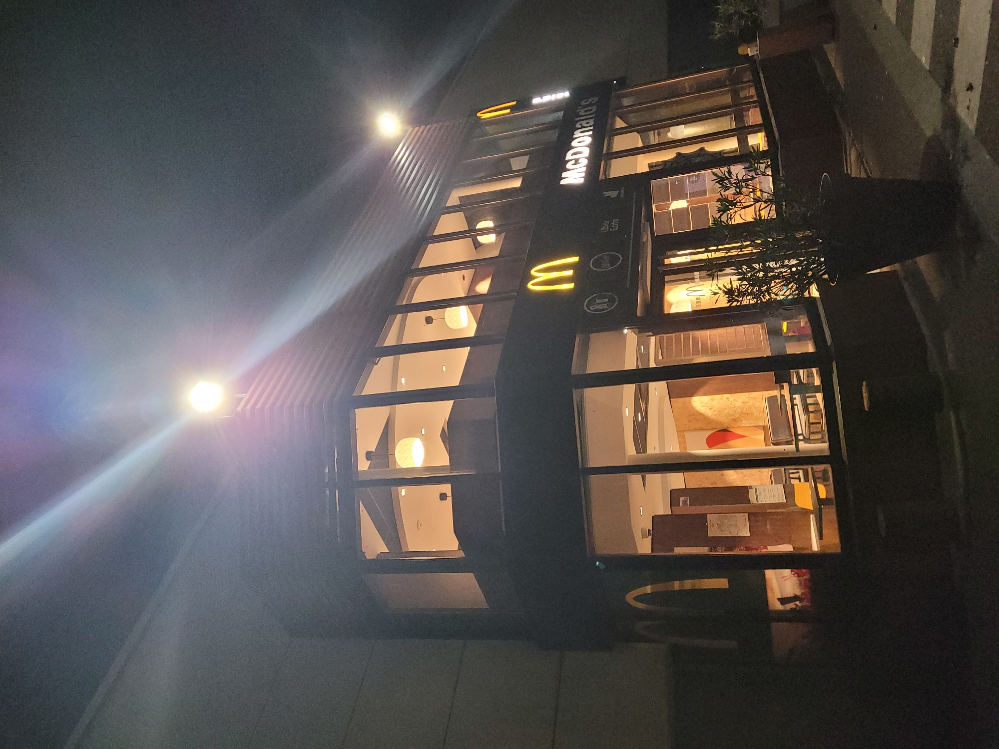 This is a nighttime exterior photograph of a McDonald's restaurant located in Givors, France. The building has a modern design with large glass windows and a prominent McDonald's logo, including illuminated golden arches and the "McDonald's" name in white lettering. The restaurant is brightly lit from within, with warm yellow lighting illuminating the interior seating areas and the drive-thru windows. A potted plant is visible in front of the entrance. The sky is dark, and the scene is illuminated by the restaurant's lights and overhead streetlights, creating lens flares and light streaks in the image. The photograph captures the building's facade and a portion of the parking lot.