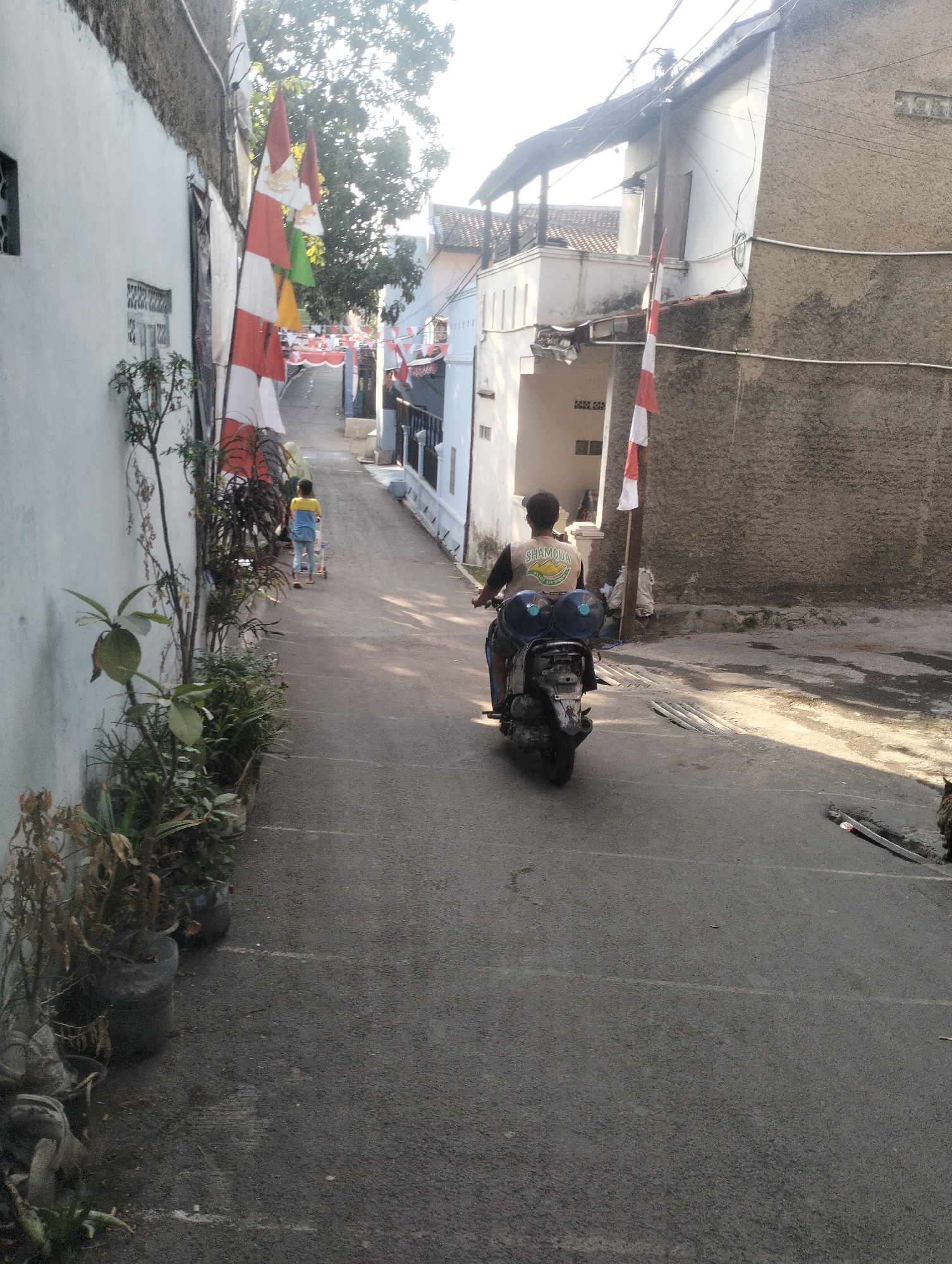 This is a street-level photograph taken in Kel. Pasir Jati, Indonesia, likely in a residential area. The scene depicts a narrow, paved street bordered by buildings and vegetation. In the foreground, on the left side of the street, are several potted plants, some appearing dry and others healthy. A white-washed wall runs along the left edge of the street. Further up the street, two children are visible, one appearing to be pushing a wheeled toy. A person is riding a scooter towards the viewer from further up the street. The scooter has two large blue water containers attached to the back. Several Indonesian flags, red and white, are mounted on poles along the street, suggesting a national holiday or celebration. The buildings on either side of the street are varied in style and condition, with some appearing to be two-story residences. The sky is bright, indicating daylight, and there are trees visible in the background. The overall impression is one of a typical Indonesian street scene, with a mix of residential life and nationalistic decorations.